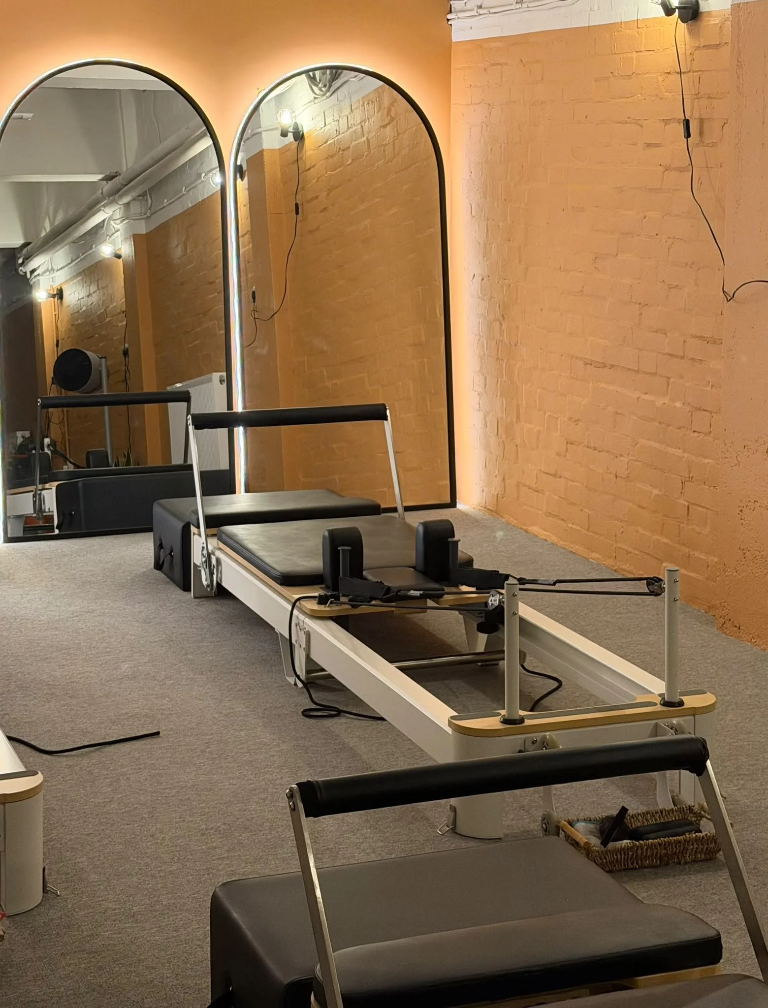 A Pilates studio with a reformer machine on a carpeted floor, orange brick walls, and large mirrors with halo lighting. The room also has a ladder barrel and a basket with yoga straps.