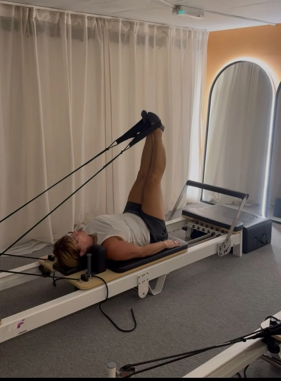A woman is lying on a specialized Pilates reformer machine in a rehab or physiotherapy room, performing an exercise with resistance bands attached to her feet.