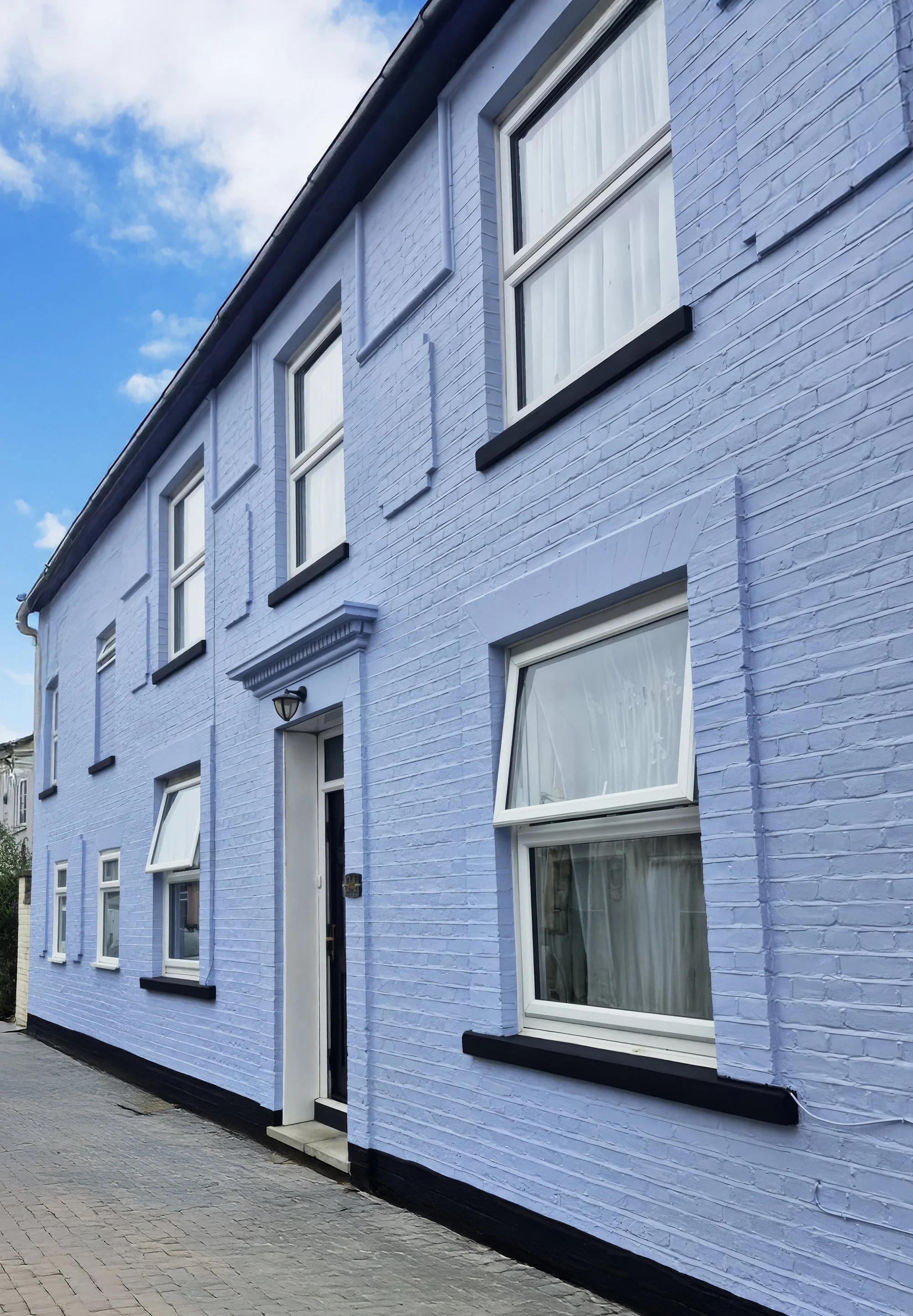 A light purple brick house with white-framed windows and a black accent trim, on a paved street, under a partly cloudy sky.