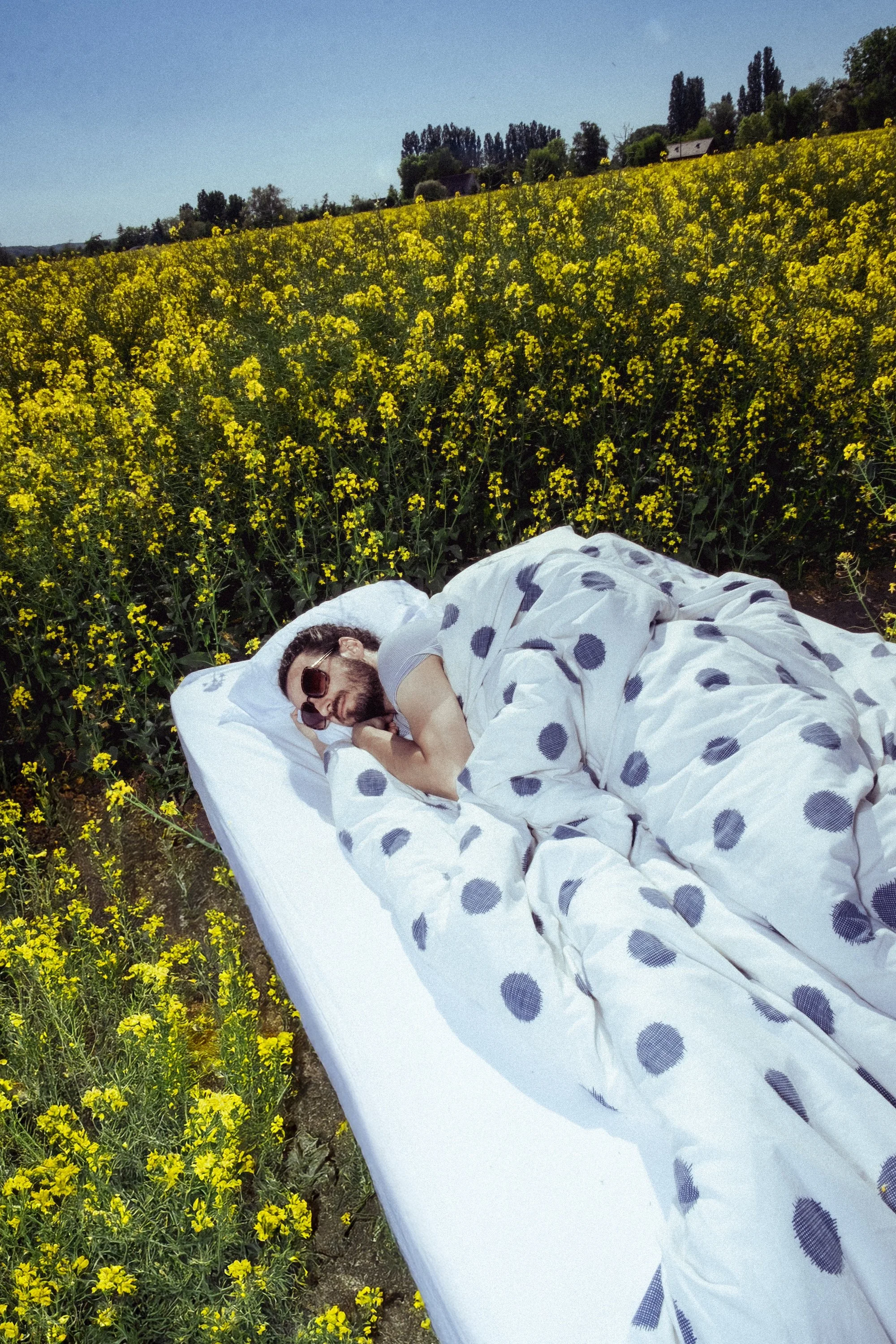 Lead singer and guitarist for Billy Bonbon, Félix Martinaud, with sunglasses sleeping on a bed outdoors in a yellow flower field on a sunny day.