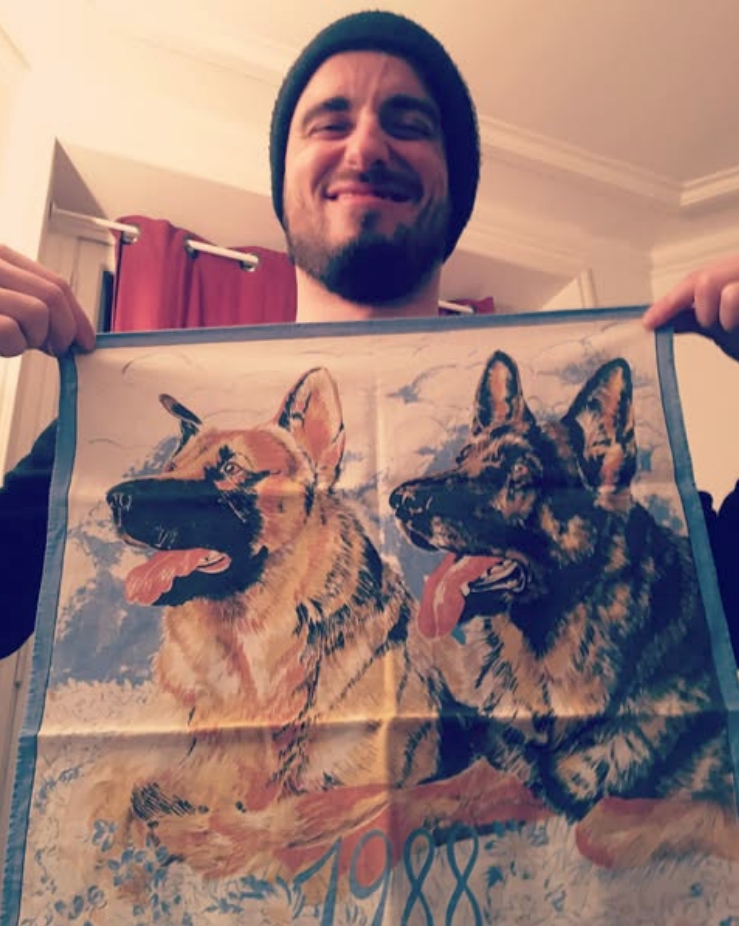 Félix Martinaud, lead singer of Billy Bonbon, wearing a black beanie and dark clothing is holding a fabric poster of two German Shepherd dogs that says '1988.' The background shows a room with red curtains and cream-colored walls.