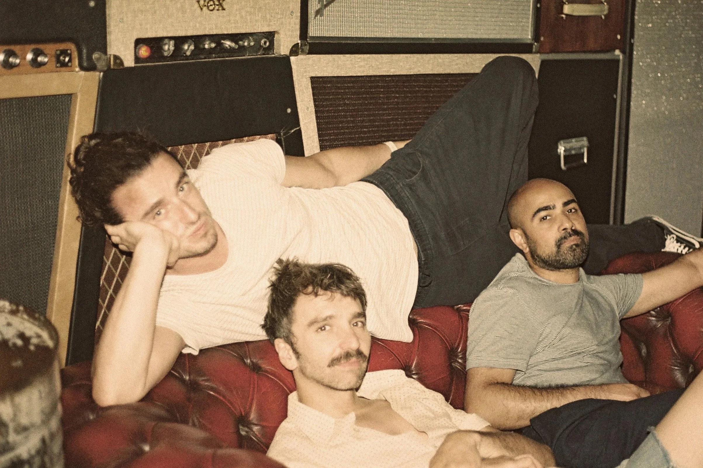 Félix Martinaud and Pablo Brunaud relaxing on a red leather couch with Jessy surrounded by vintage guitar amplifiers in a music studio.