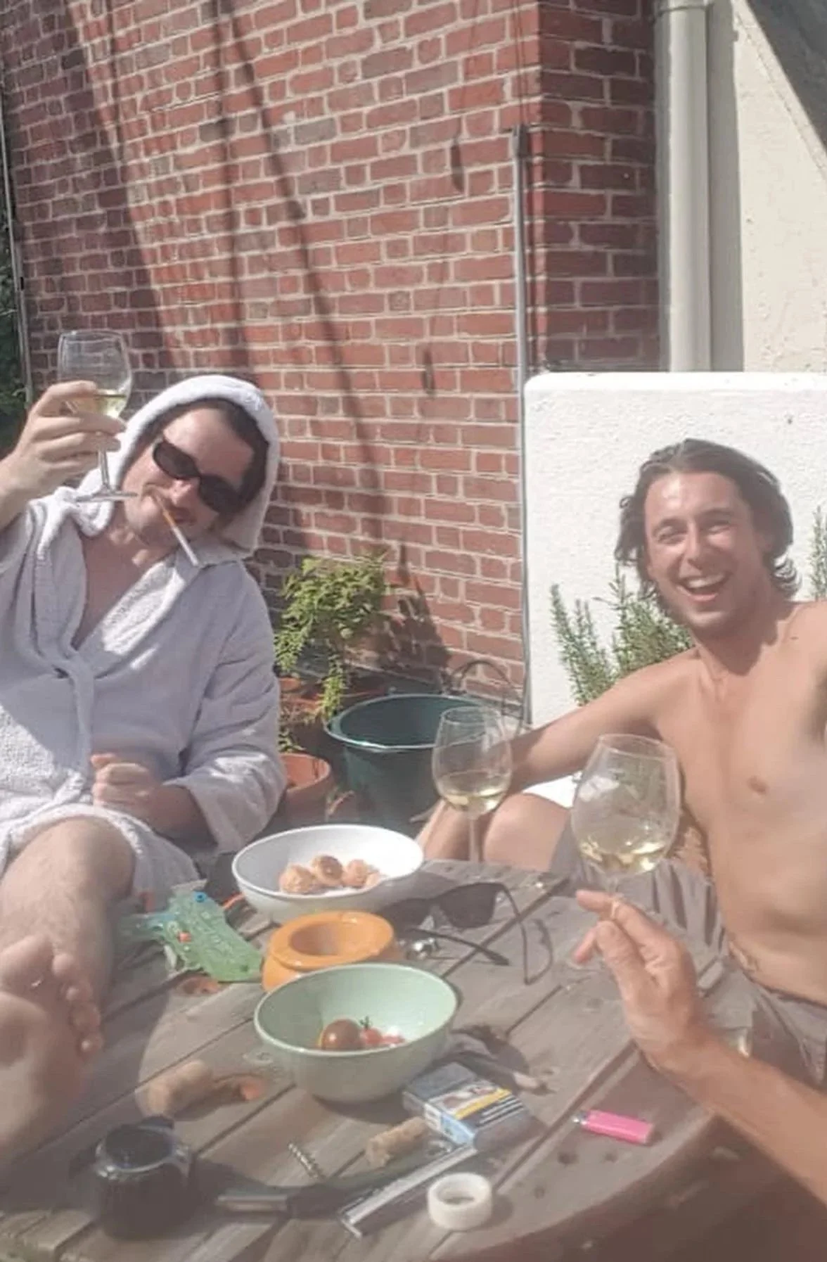Félix Martinaud, lead singer of Billy Bonbon, sitting with a friend outdoors at a table, one wearing sunglasses and a bathrobe, the other shirtless, smiling, both holding wine glasses, with food and objects on the table, in front of a brick wall.