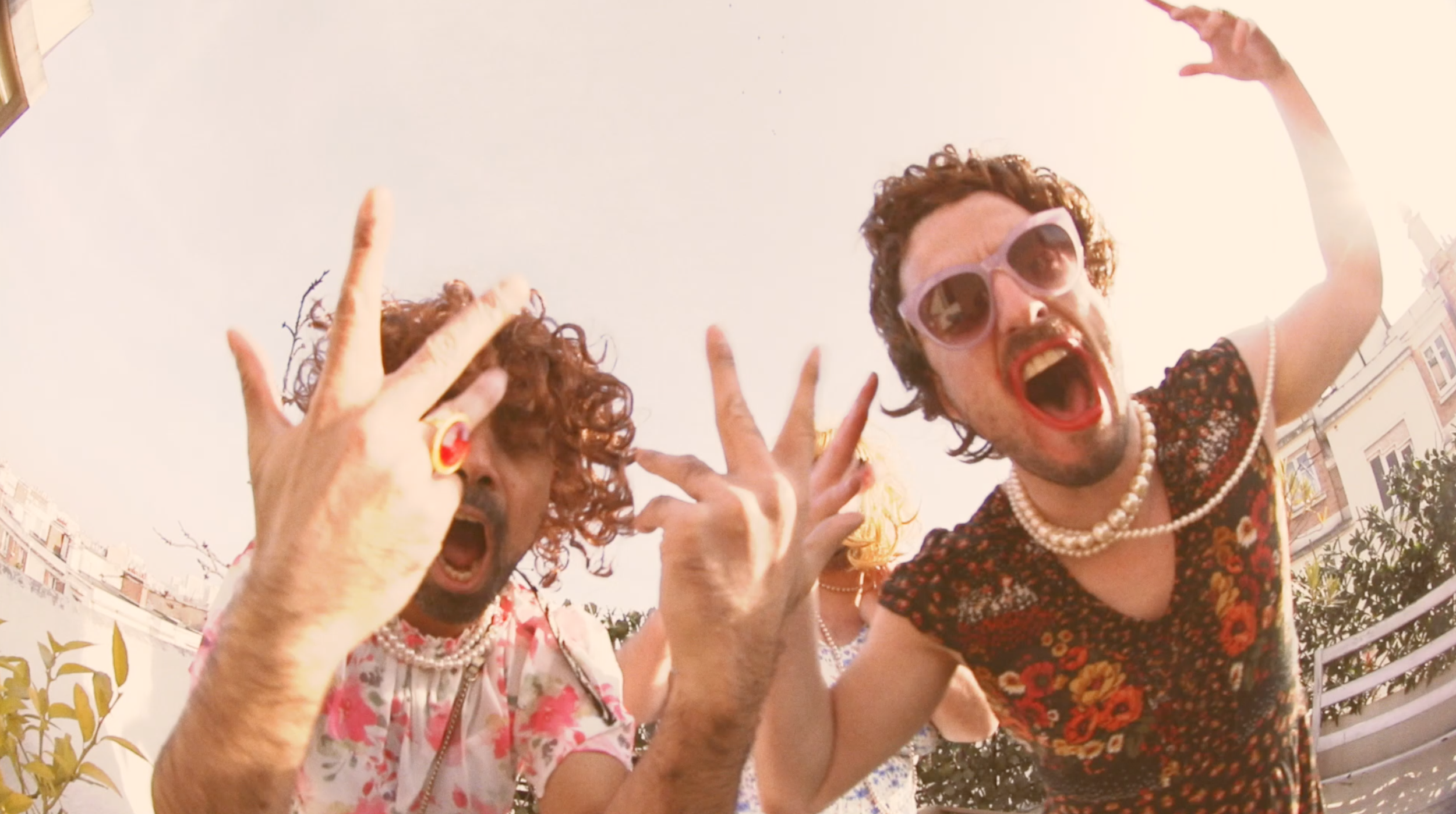Pablo Brunaud and Félix Martinaud on set for their music video, Billy Bonbon, dressed in colorful, retro-style clothing with pearl necklaces, sunglasses, and bold makeup pose playfully outdoors against a bright Paris sky and city buildings.