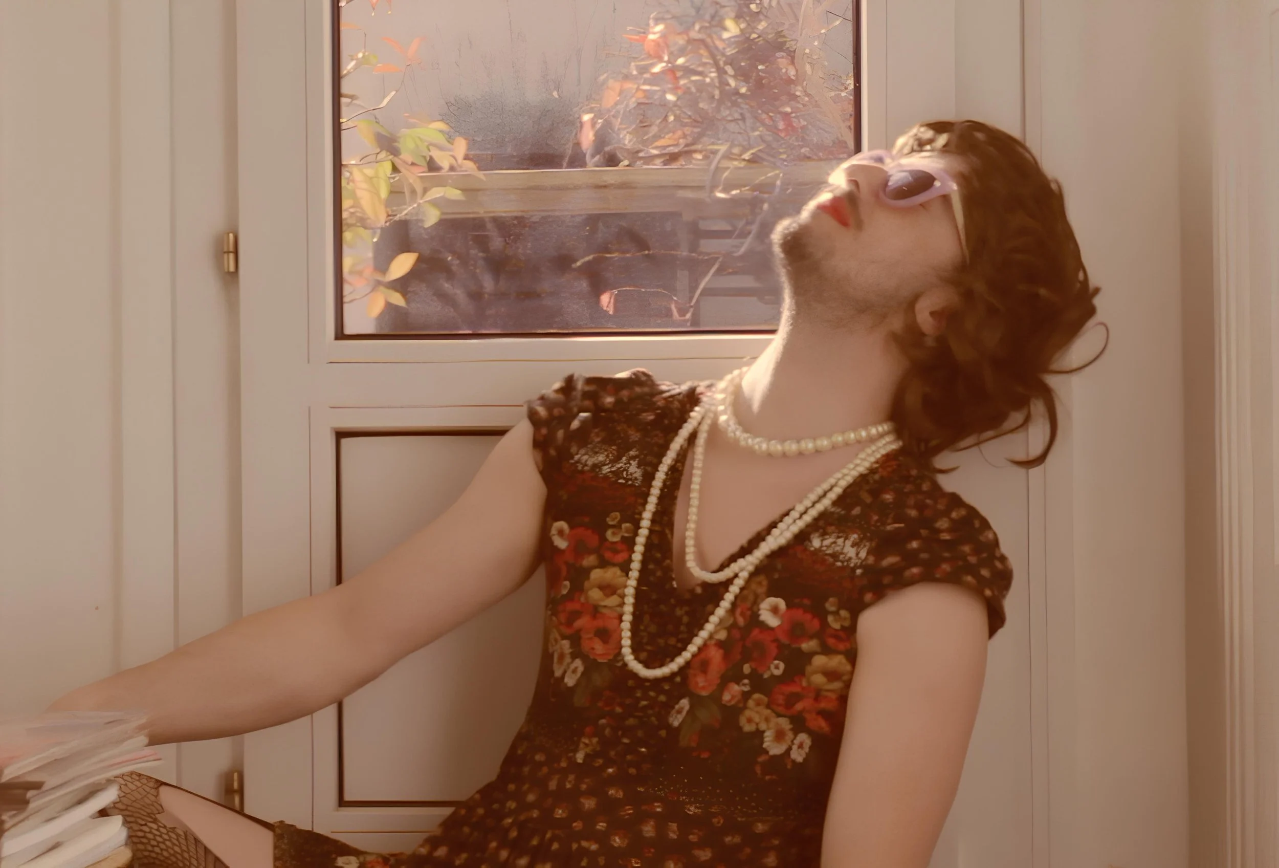 Félix Martinaud of Billy Bonbon with curly hair wearing pink sunglasses, a flower-patterned dress, and pearl necklaces, sitting by a window with closed wooden shutters, looking upward with a relaxed expression.