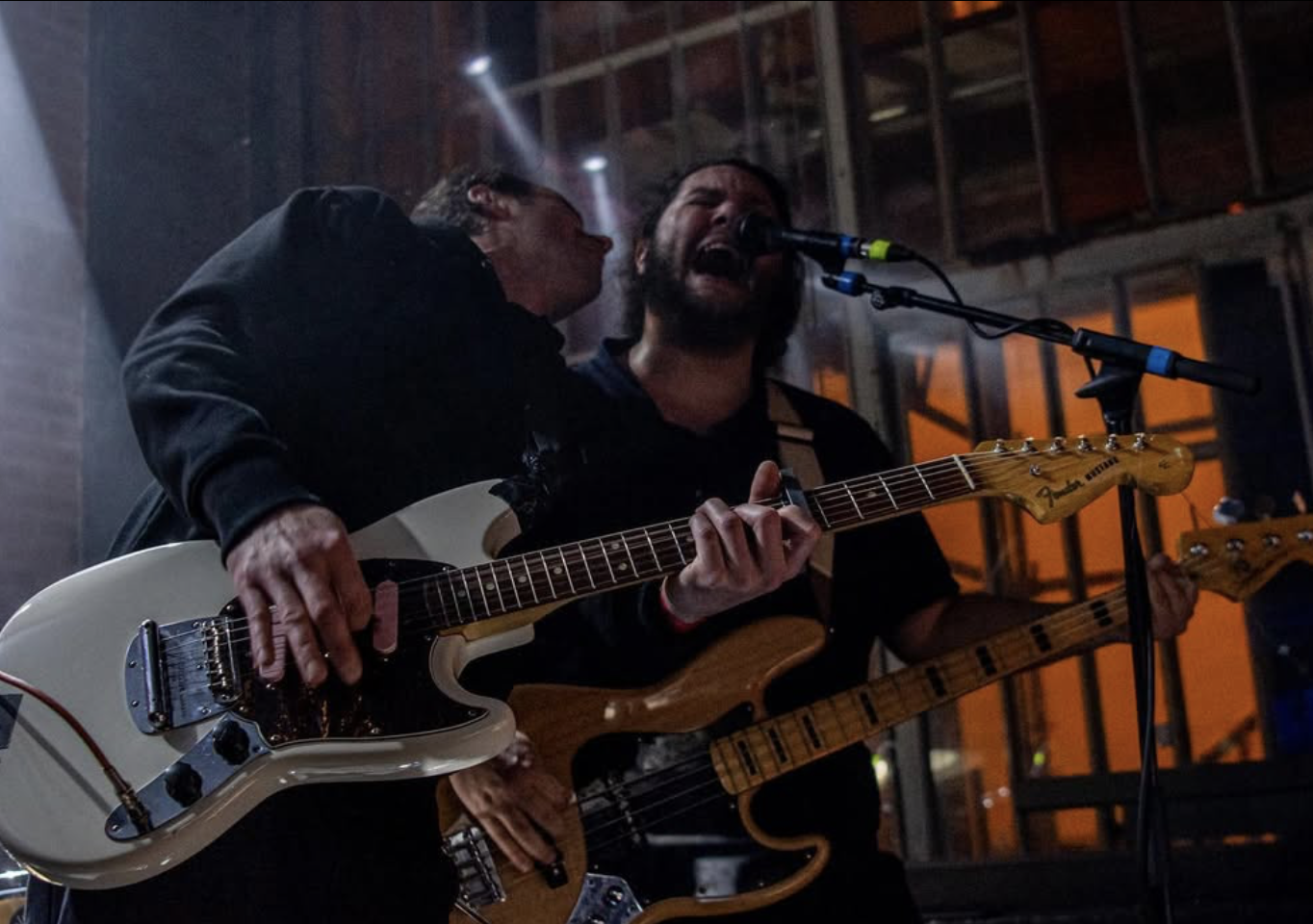 Two musicians performing on stage, one singing into a microphone and playing electric guitar, the other playing bass guitar in a dimly lit venue.
