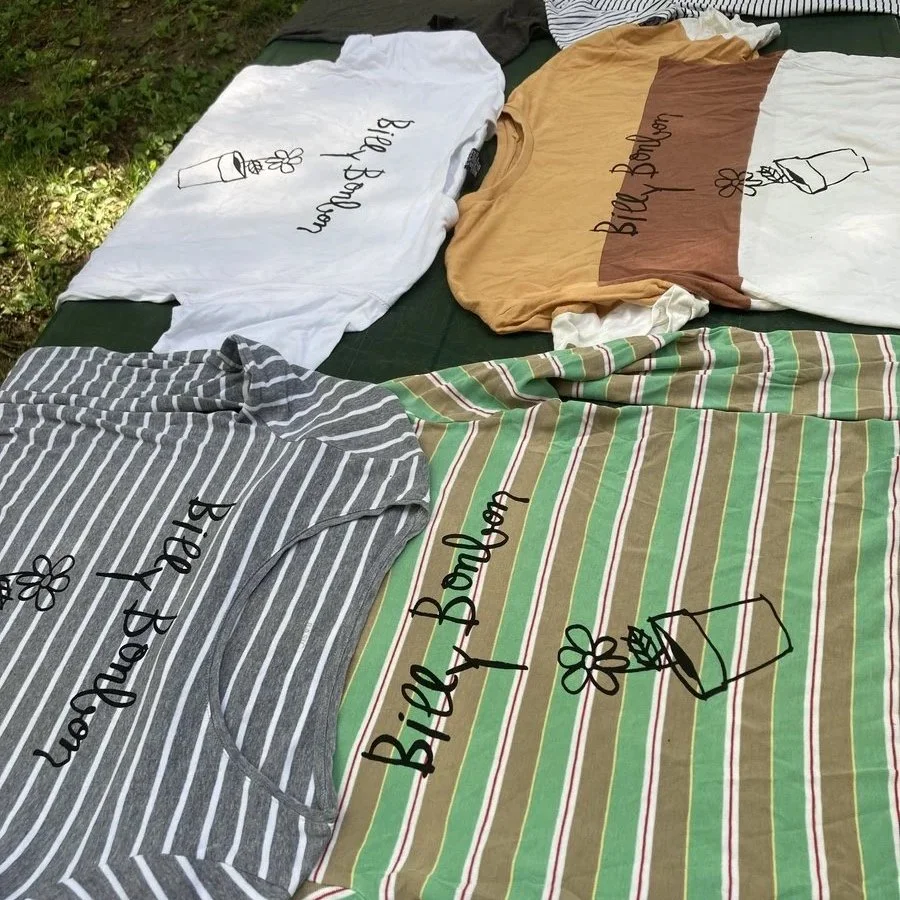 Four Billy Bonbon band branded T-shirts with similar design laid out on a bed or outdoor surface. The T-shirts have the text 'Billy' and a simple line drawing of a flower in a pot, each in different colors and patterns.