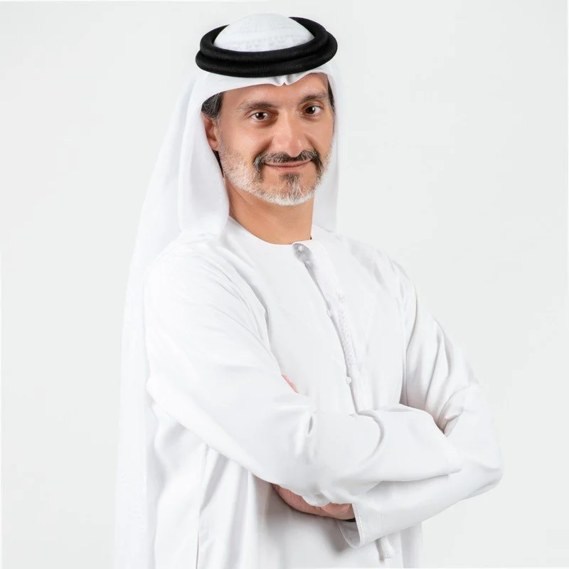 A middle-aged man wearing traditional Middle Eastern attire, including a white kandura, a white ghutra, and a black agal, smiling with arms crossed against a plain white background.