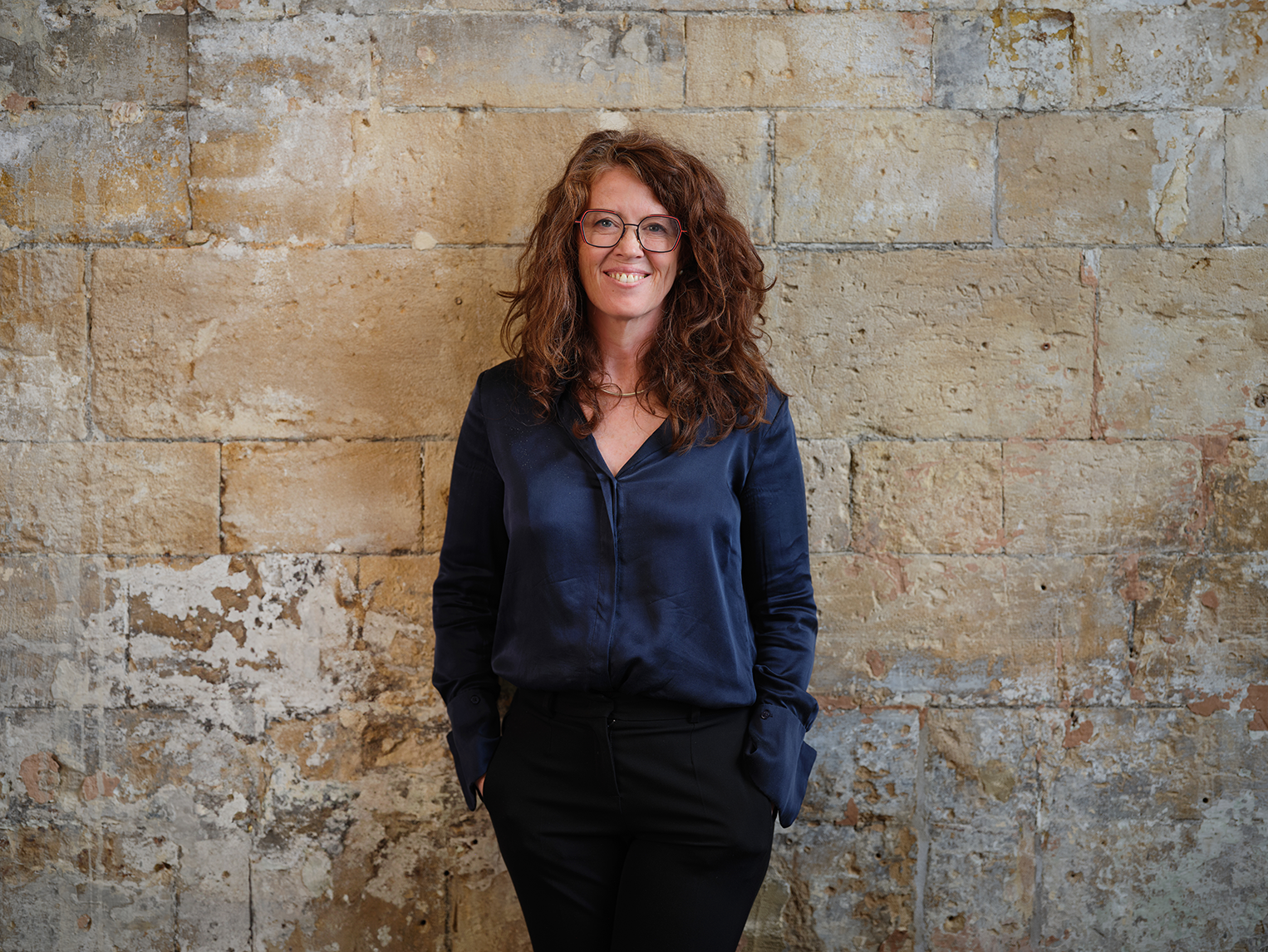 A woman with curly red hair, glasses, wearing a navy blue blouse and black pants, standing against a textured stone wall, smiling at the camera.