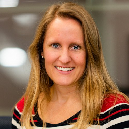 A woman with long blonde hair and blue eyes smiling, wearing a red, black, and white striped top, in an indoor setting with blurred background.