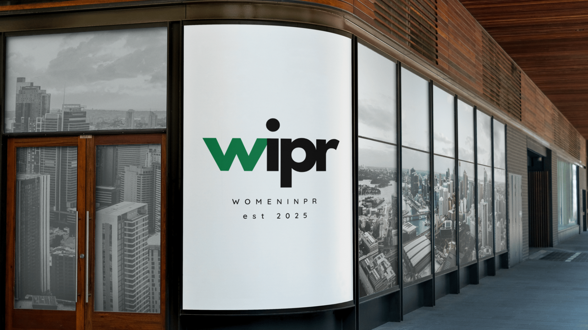 Signboard for WOMAN IN PR with logo reading 'wipr' and text 'WOMENINPR est 2025' on a cityscape window background.
