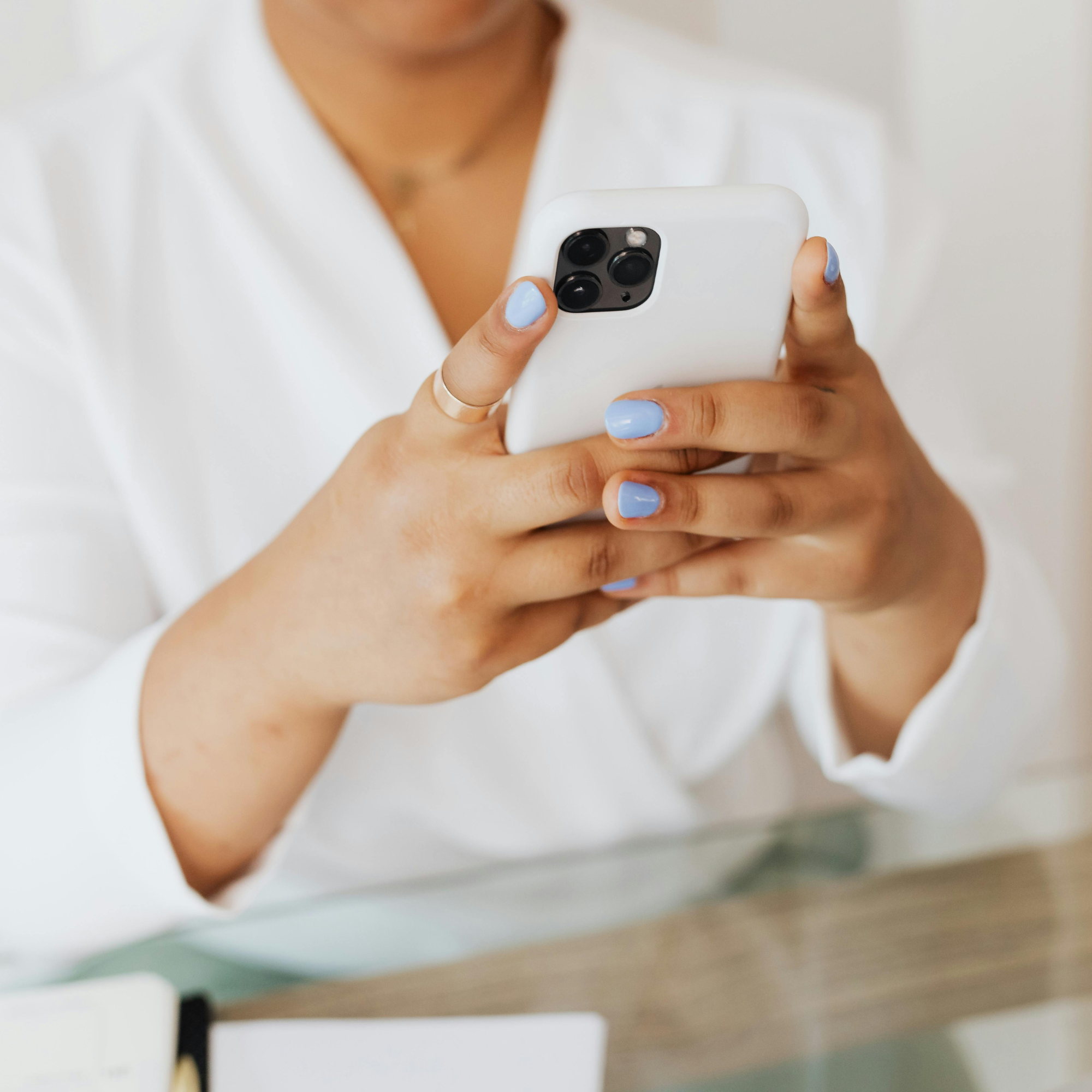 Person with light blue painted nails holding a white smartphone while sitting at a table.