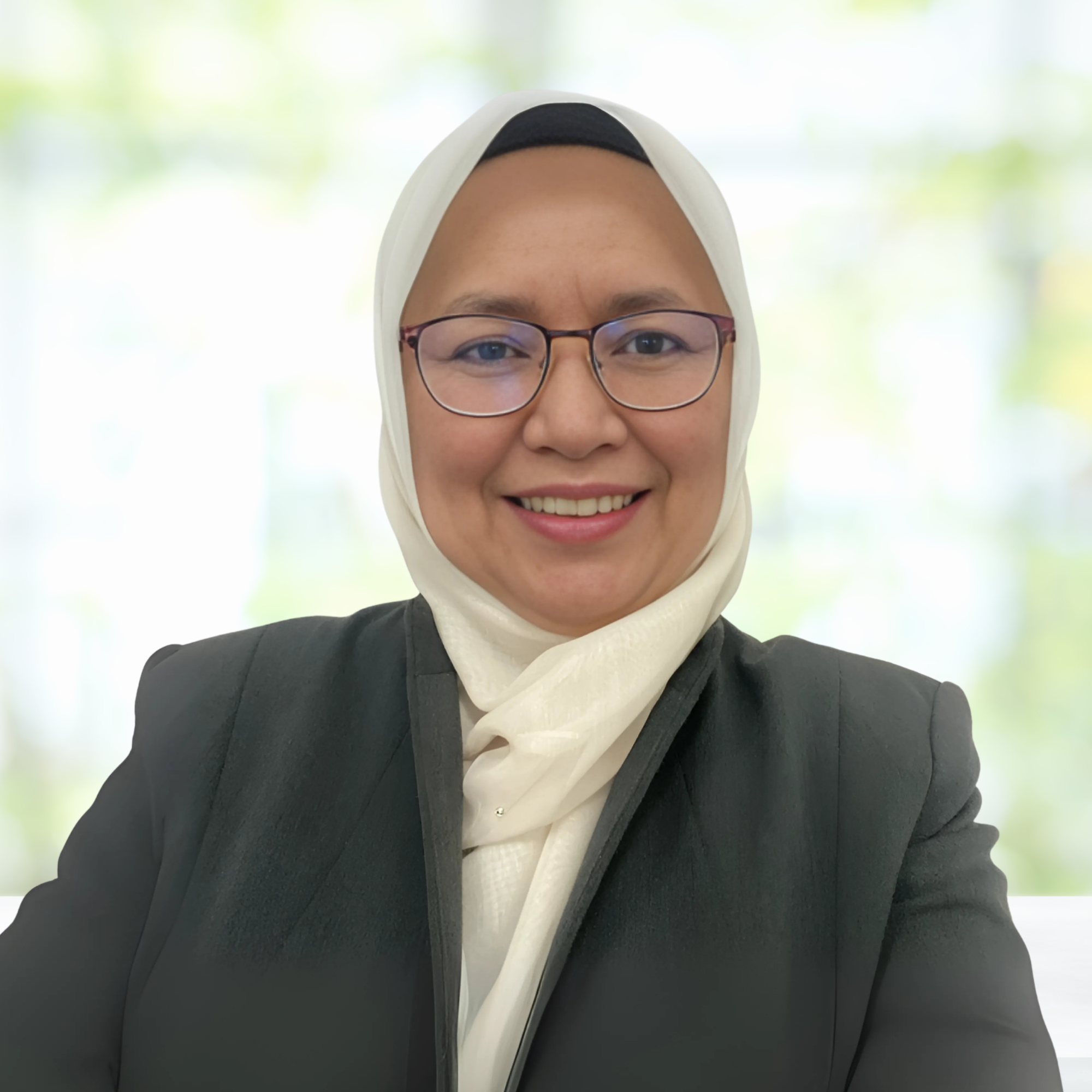 A woman wearing a white hijab and glasses, smiling at the camera with a blurred light-colored background.