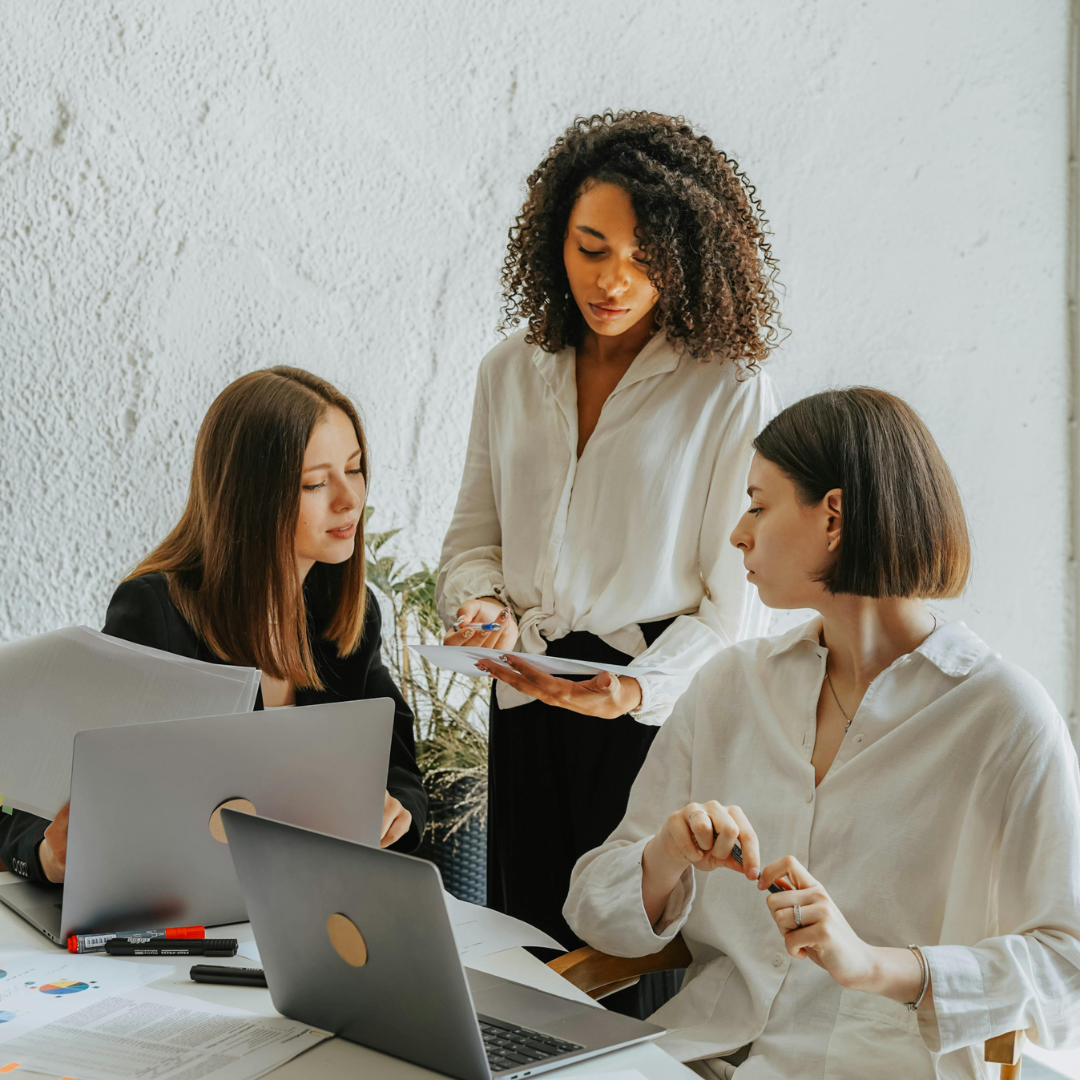 Three women in an office working together, two seated at a desk with laptops and documents, one standing with a notepad.