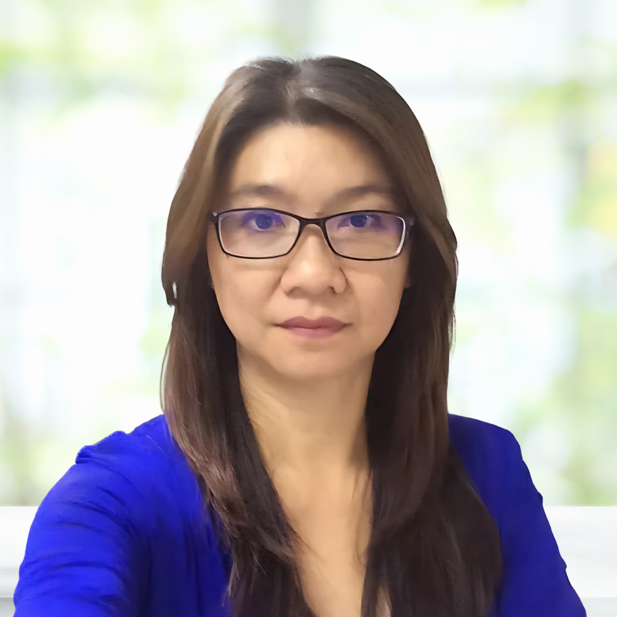 Portrait of a woman with long dark hair and glasses, wearing a blue top, standing in front of a bright, blurred background.