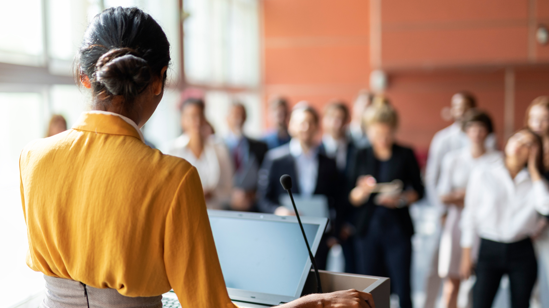 A woman in a yellow blazer speaking at a podium with a laptop, addressing a group of people in a conference room.
