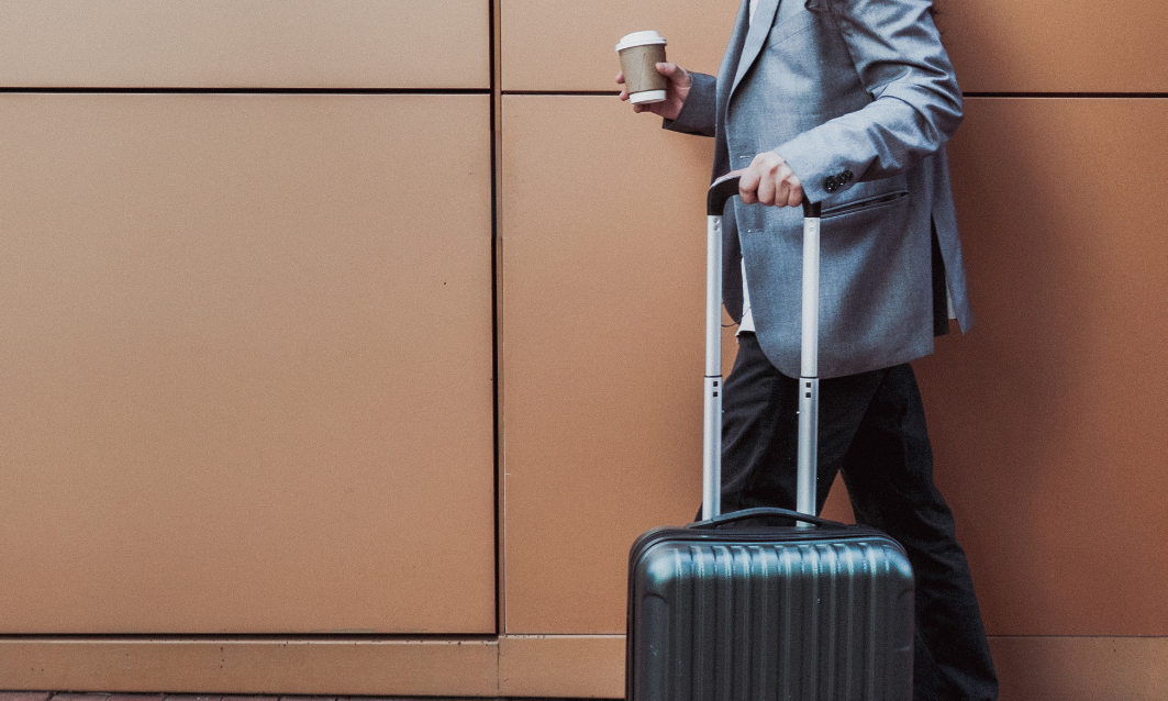 A person in a gray blazer holding a coffee cup and pulling a black rolling suitcase against a tan wall.