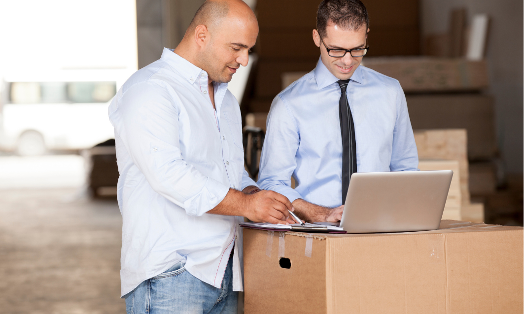 Two men in office shirts and glasses working together on a laptop and clipboard in a warehouse.
