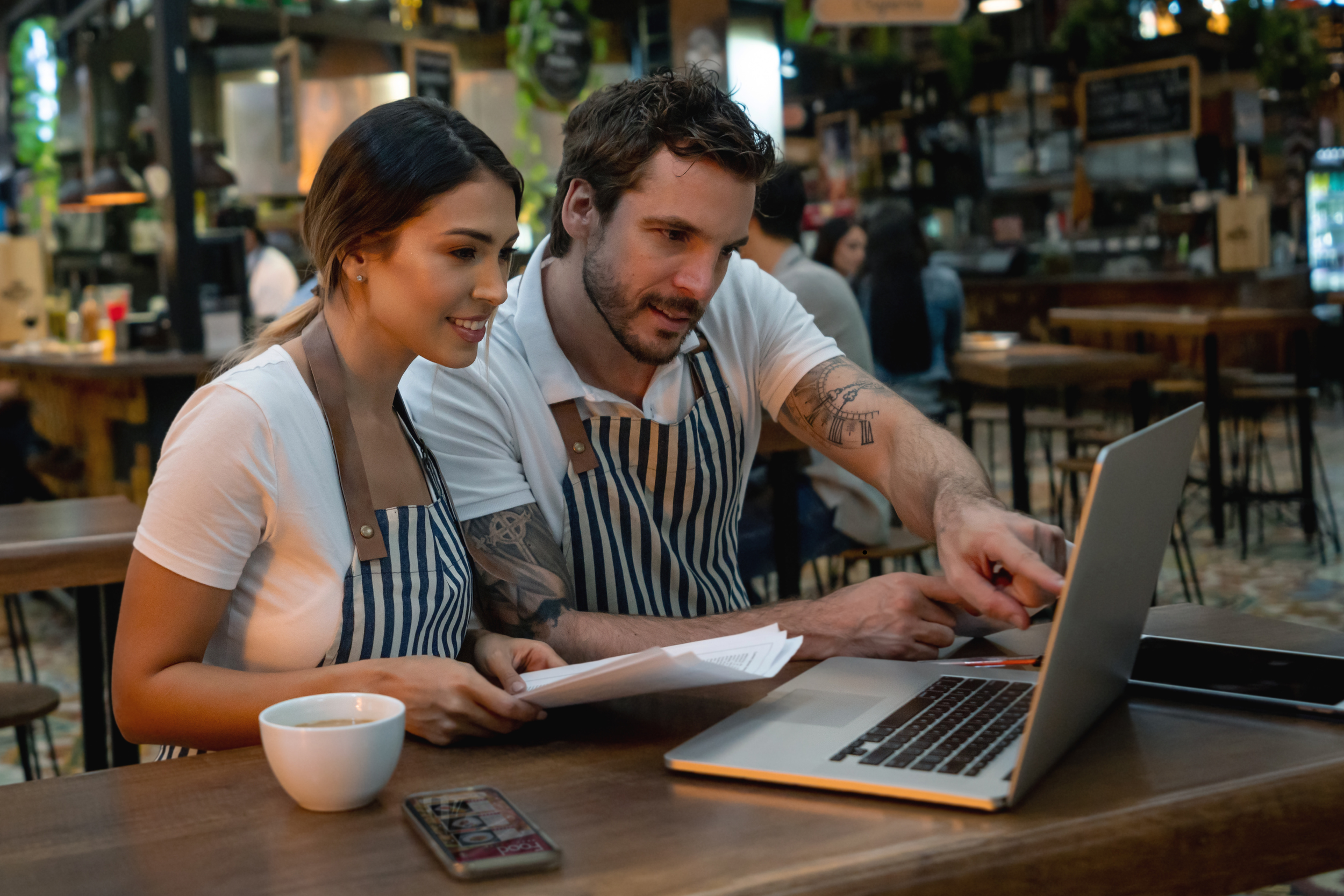 Two restaurant workers, a woman and a man, wearing striped aprons, are looking at a laptop screen and discussing something while seated at a wooden table.