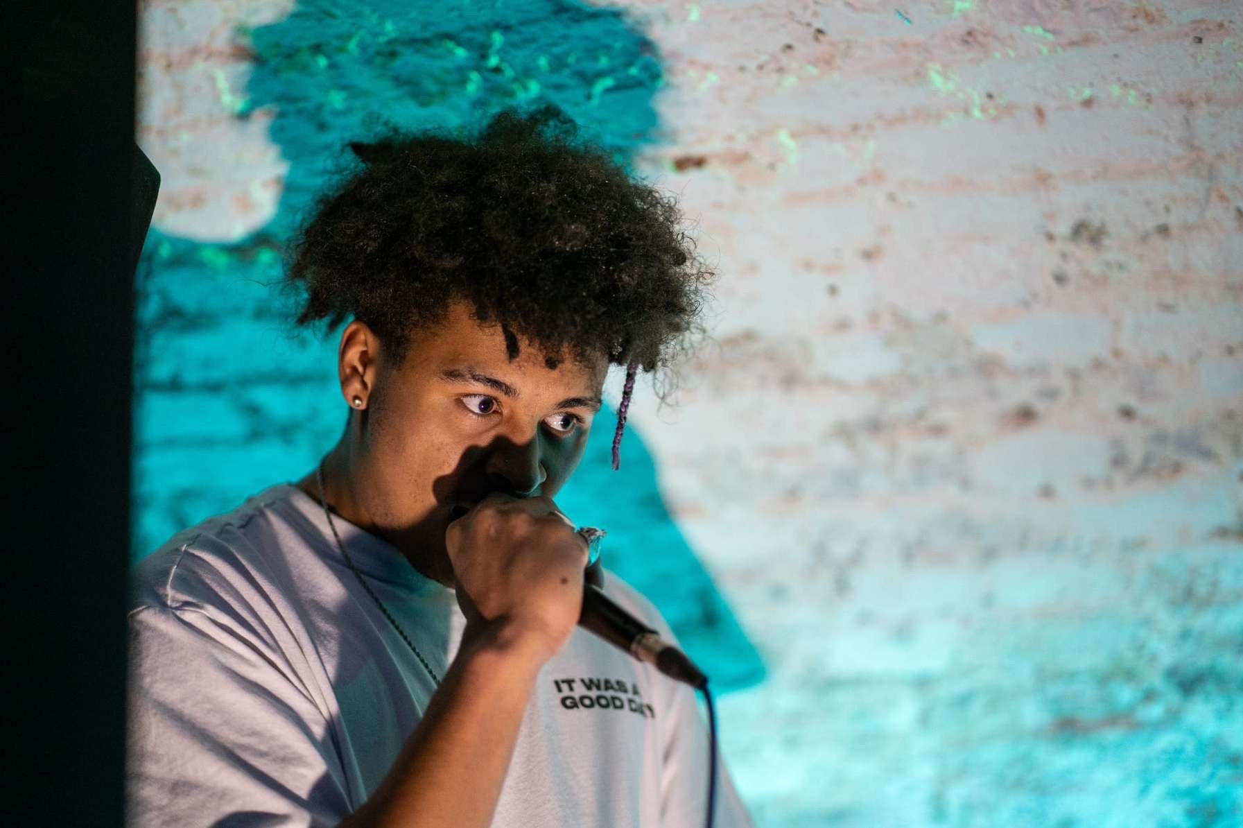 A person with curly hair holding a microphone near their mouth, standing in front of a colorful, textured background.