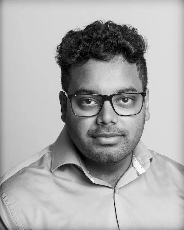 Black and white portrait of a young man with curly hair wearing glasses and a collared shirt, looking directly at the camera.