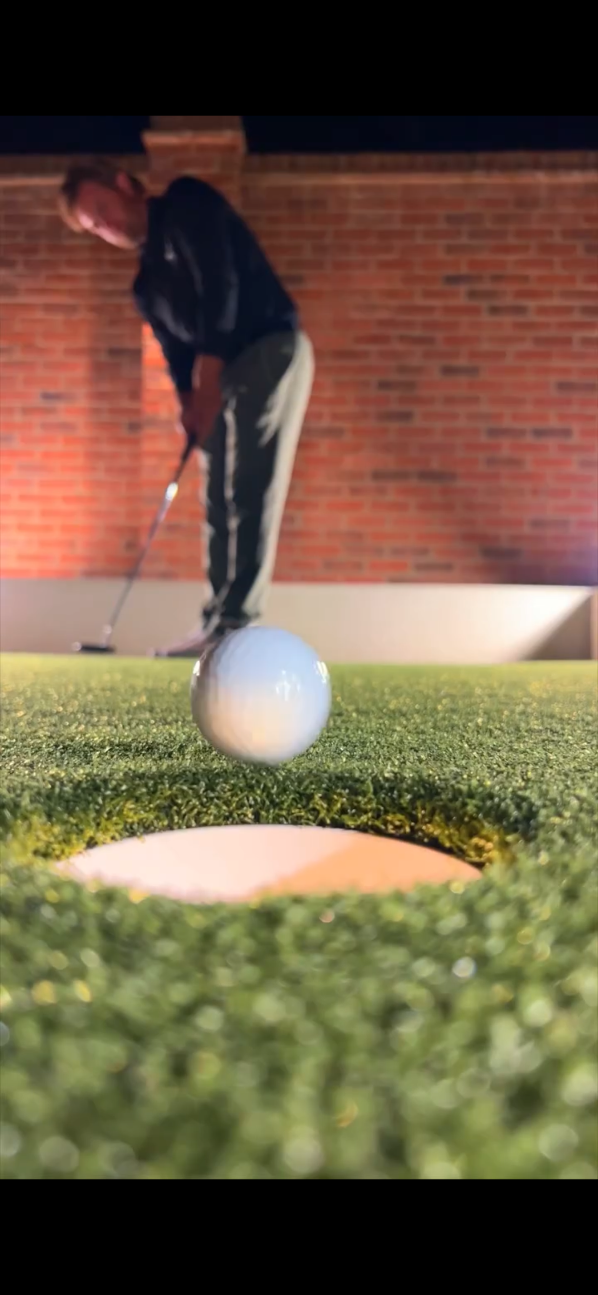 A person playing mini golf at night, preparing to hit a white golf ball towards the hole on a green artificial turf with a brick wall in the background. Home putting green