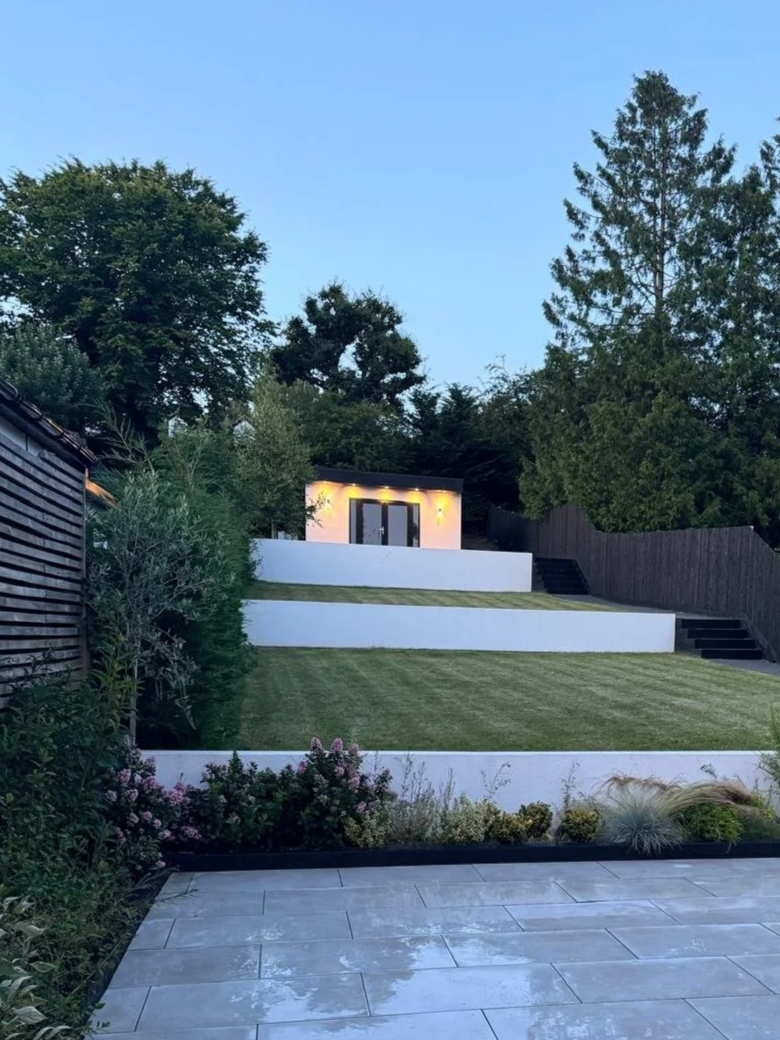 A modern backyard with tiered white retaining walls, a well-manicured lawn, an outdoor shed with lighting, and surrounded by trees and plants, with a tiled patio in the foreground.