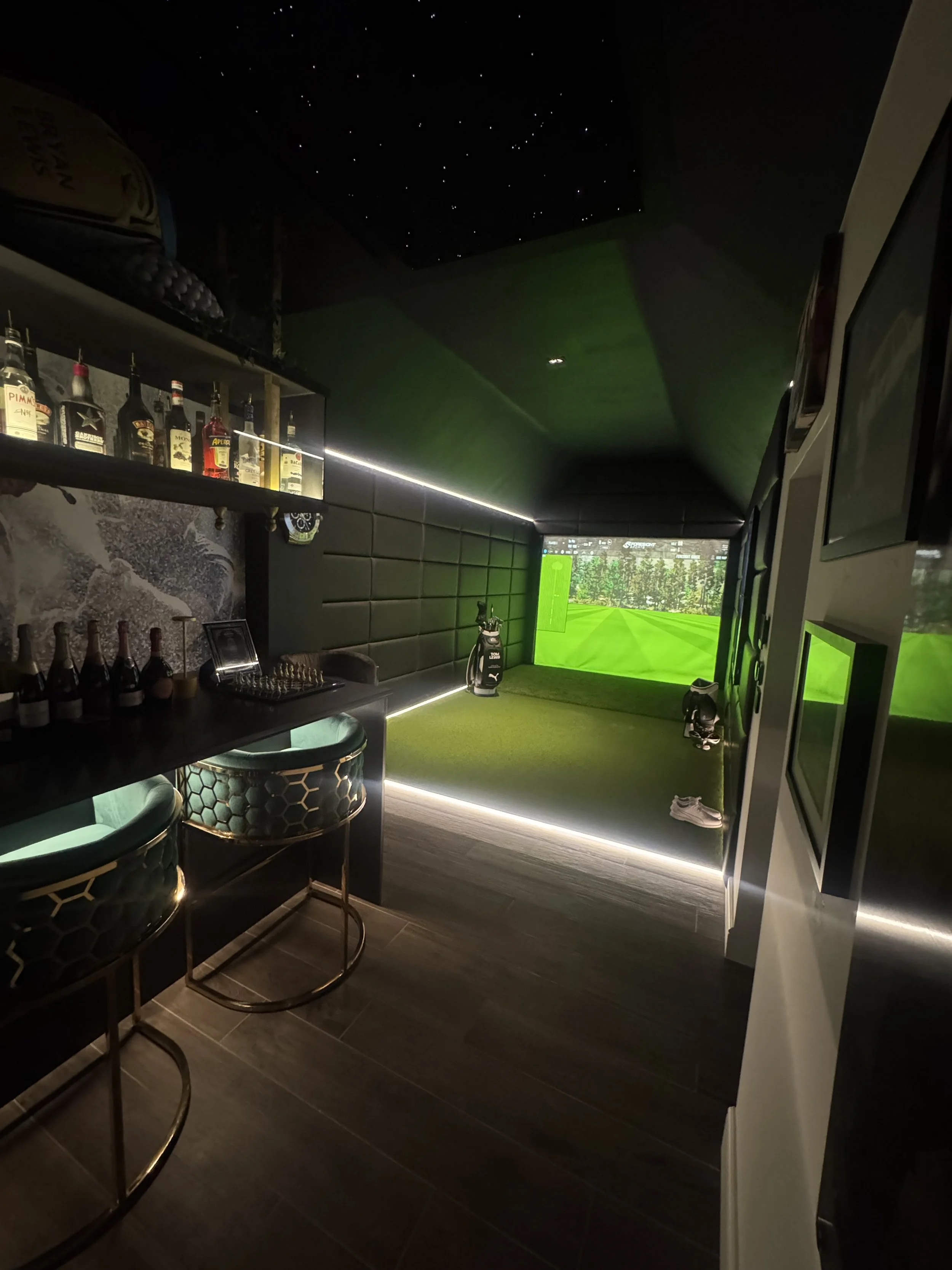 Indoor golf simulation room with a large green screen displaying a golf course, golf clubs, shoes, and golf balls, with a bar area and chairs. Foresight Golf Simulator