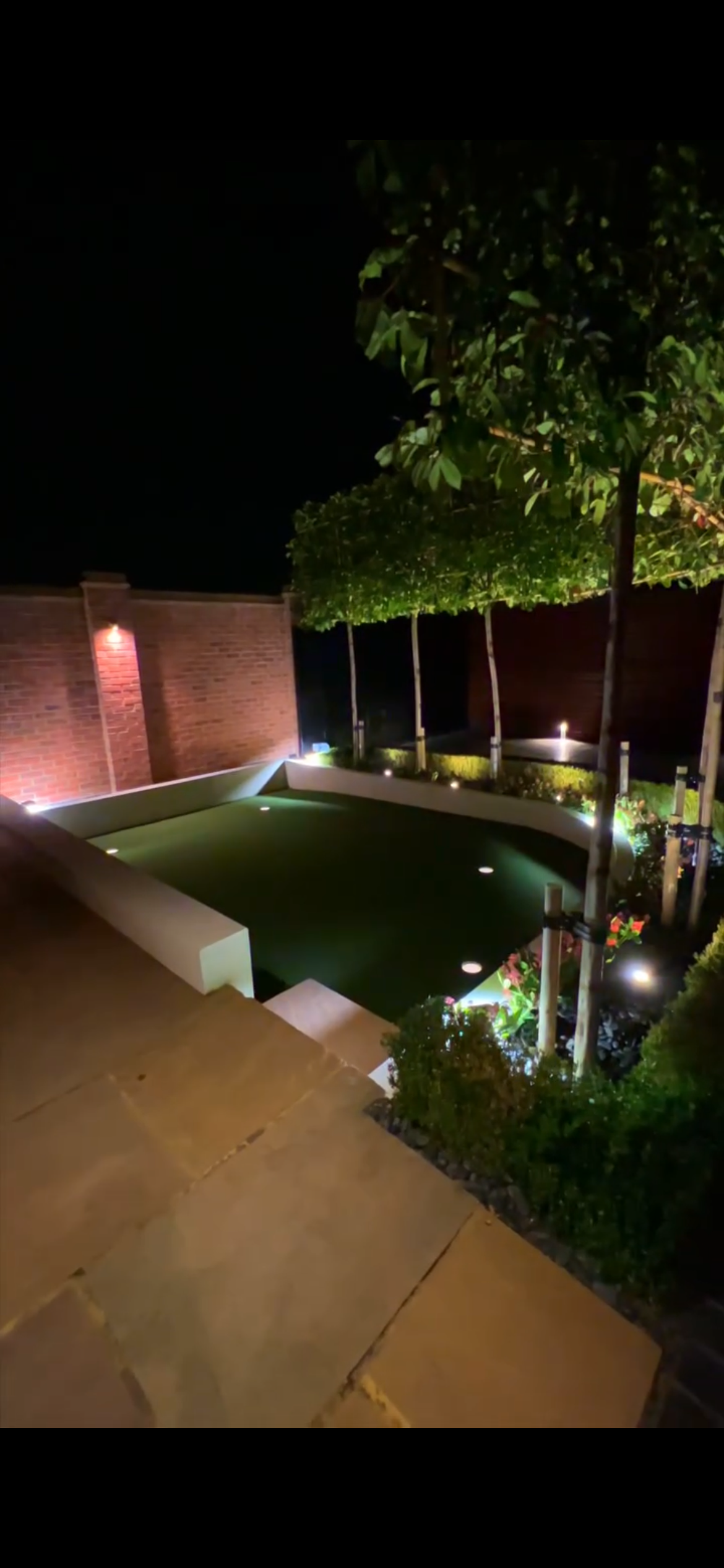 Night view of a backyard with a putting green surrounded by potted plants and trees, with a brick wall and outdoor lighting in the background.