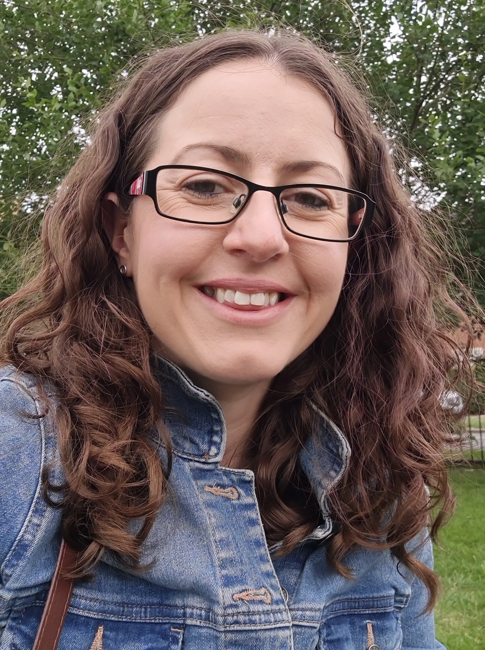 Head-and-shoulders portrait of a smiling person with wavy brown hair and glasses, wearing a blue denim jacket, photographed outdoors with trees in the background