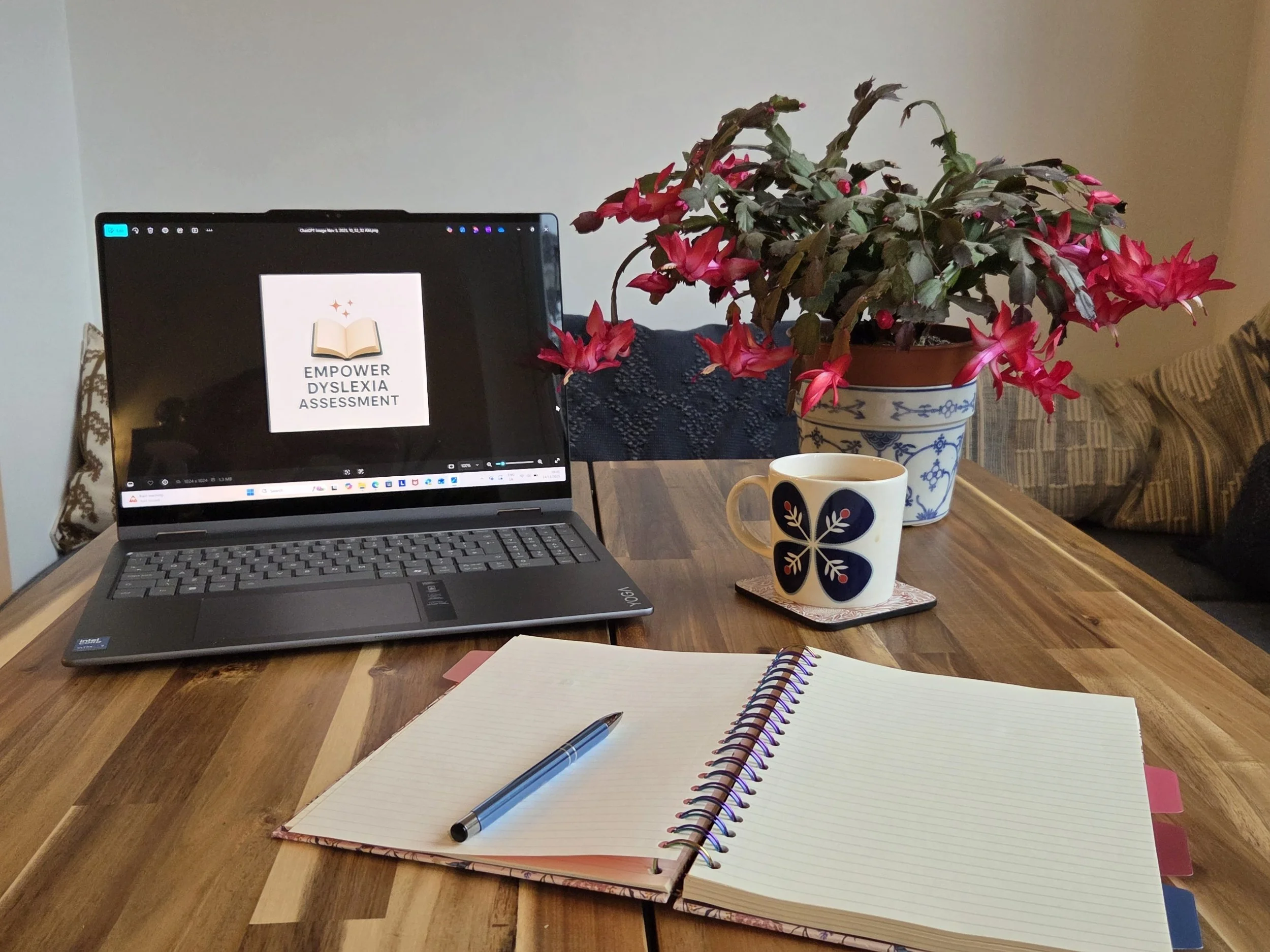 A wooden table with a laptop displaying 'Empower Dyslexia Assessment,' an open notebook with a pen, a mug on a coaster, a large potted plant with pink flowers, and a sofa with striped cushions in the background.