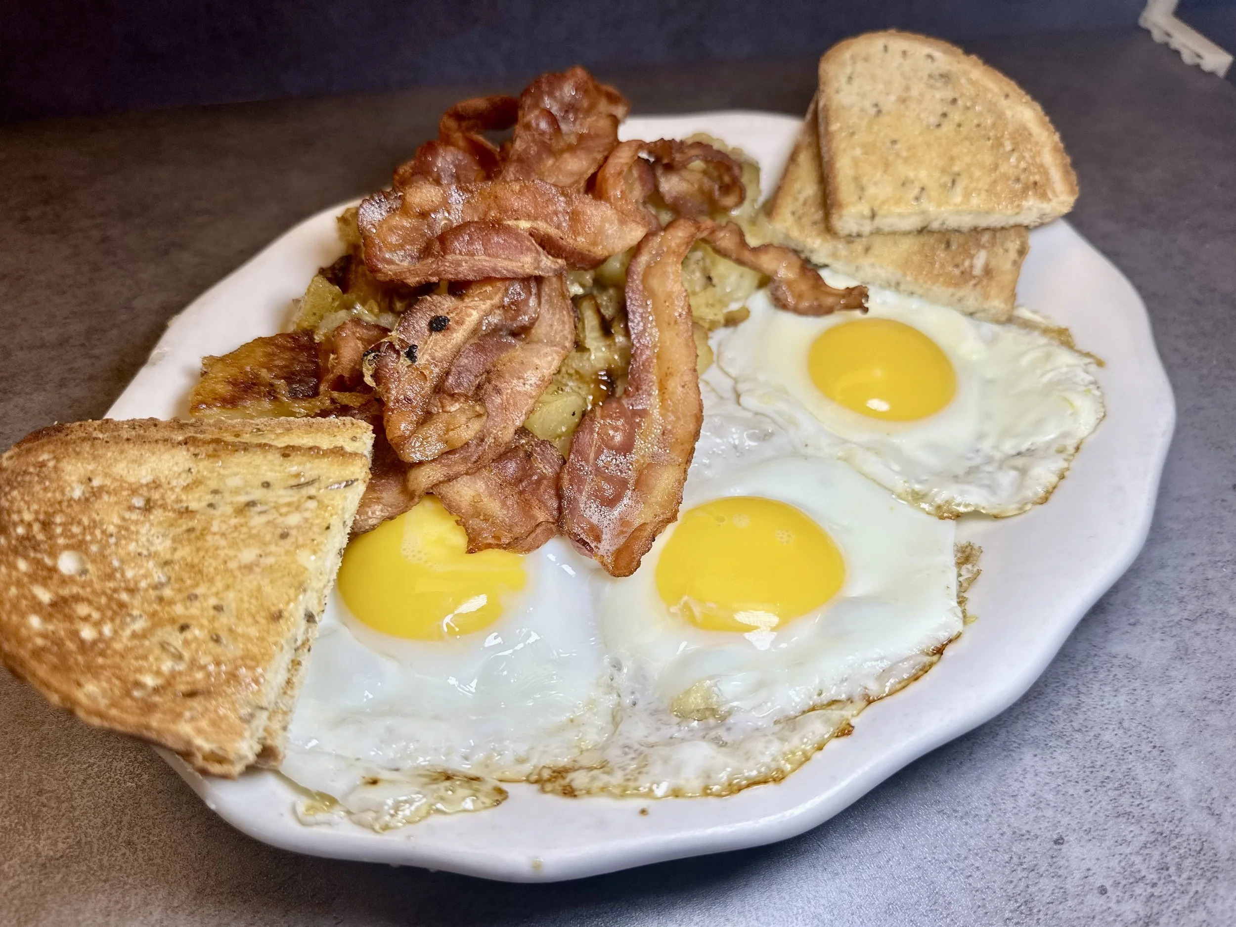 Breakfast plate with three fried eggs, crispy bacon, toasted bread slices, and potato wedges on a white dish.