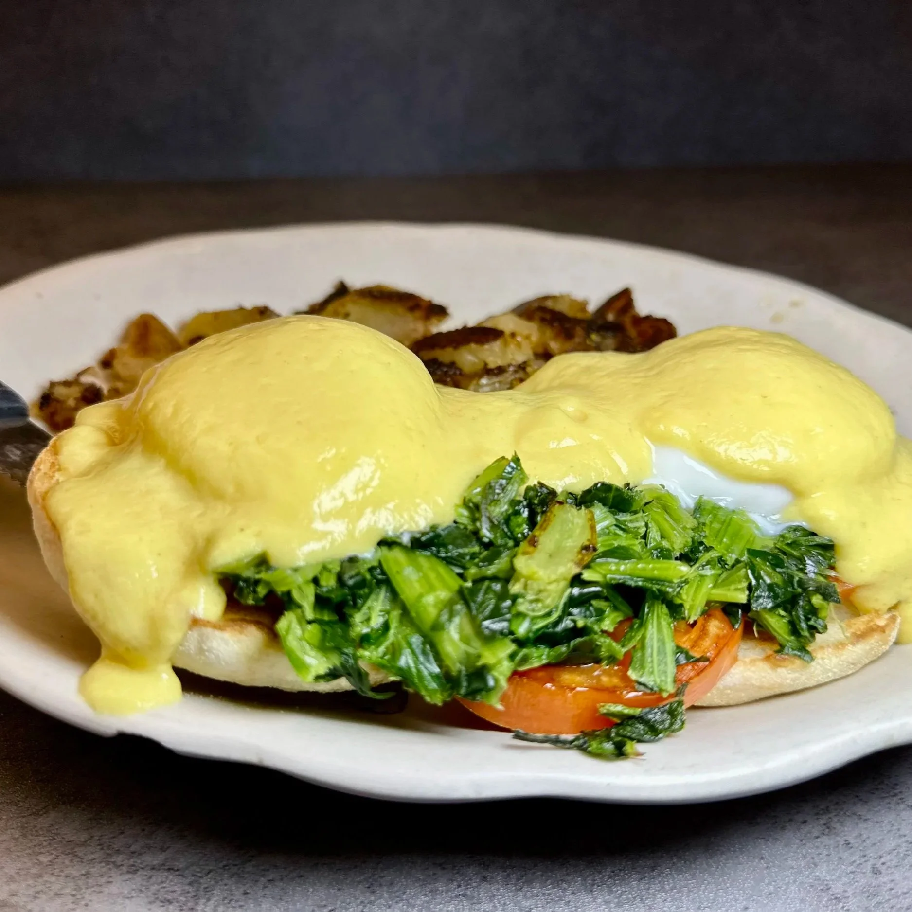 Eggs Benedict with hollandaise sauce, on a toasted English muffin with tomato, spinach, and bacon in the background.