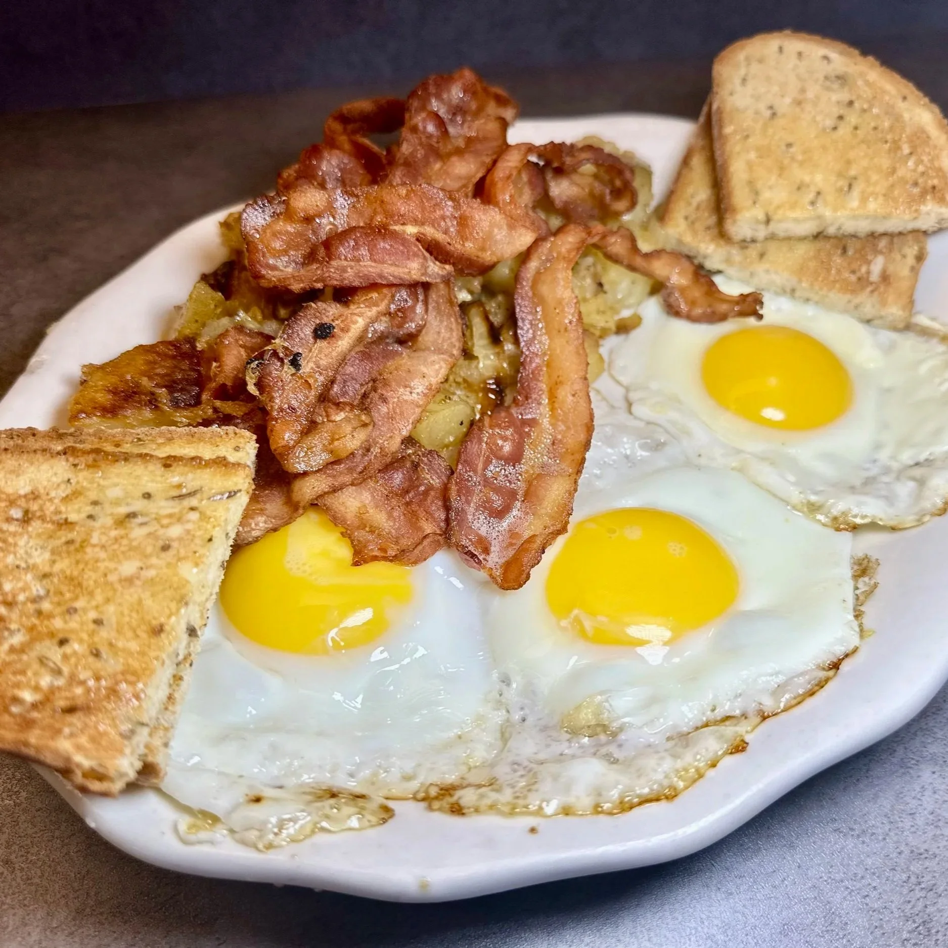 A plate of breakfast food with three fried eggs, crispy bacon, and toasted bread triangles.