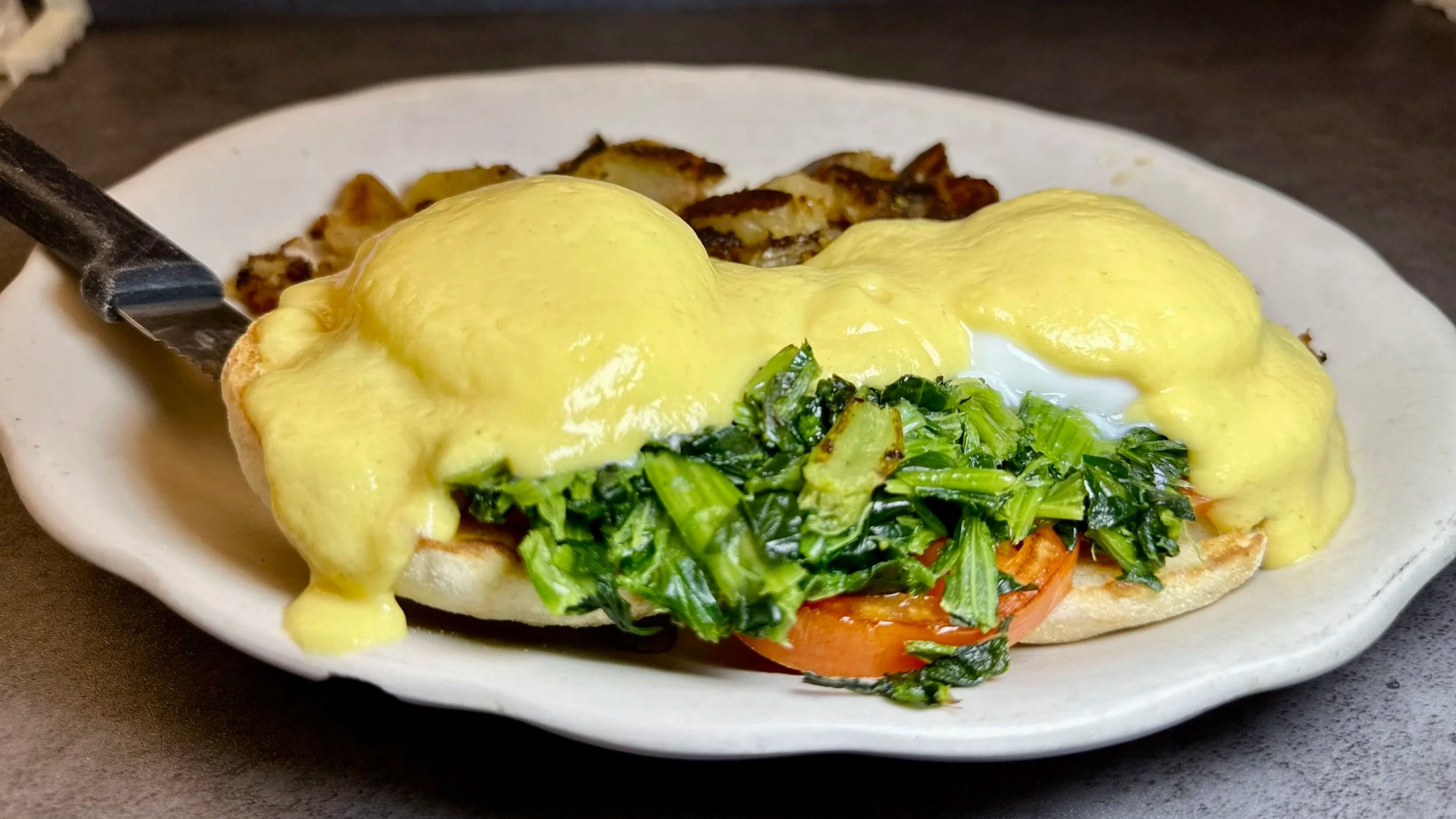 A plate with eggs benedict topped with hollandaise sauce, served with sautéed greens and tomato slices on a toasted bun.