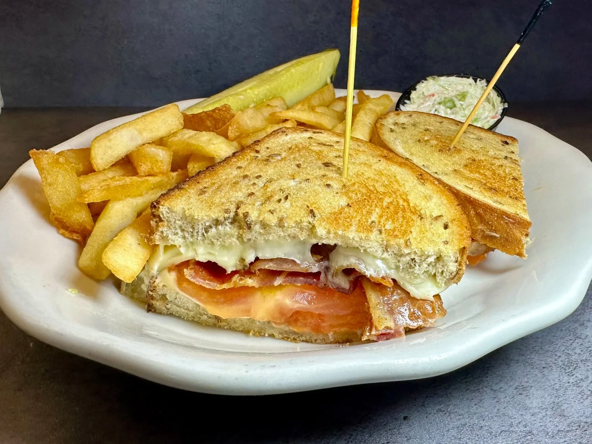Grilled cheese sandwich with tomato, served with French fries, cole slaw, and pickles on a white plate.