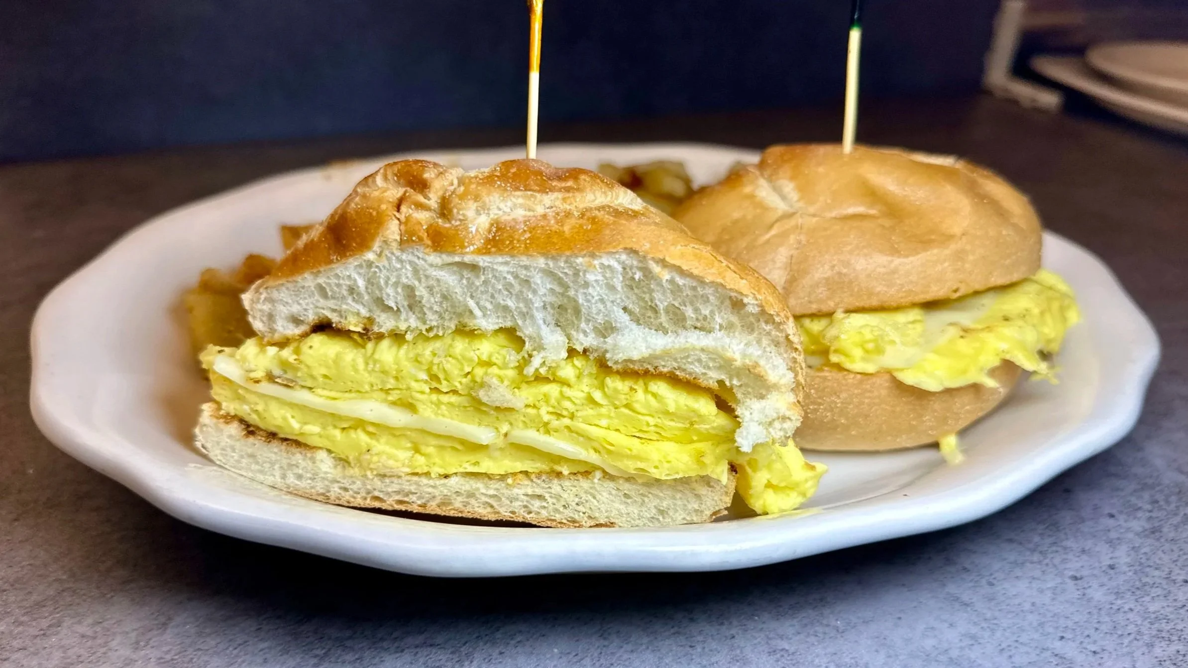 A plate with a sliced breakfast sandwich featuring scrambled eggs and a sandwich with scrambled eggs and a bun, both secured with toothpicks.