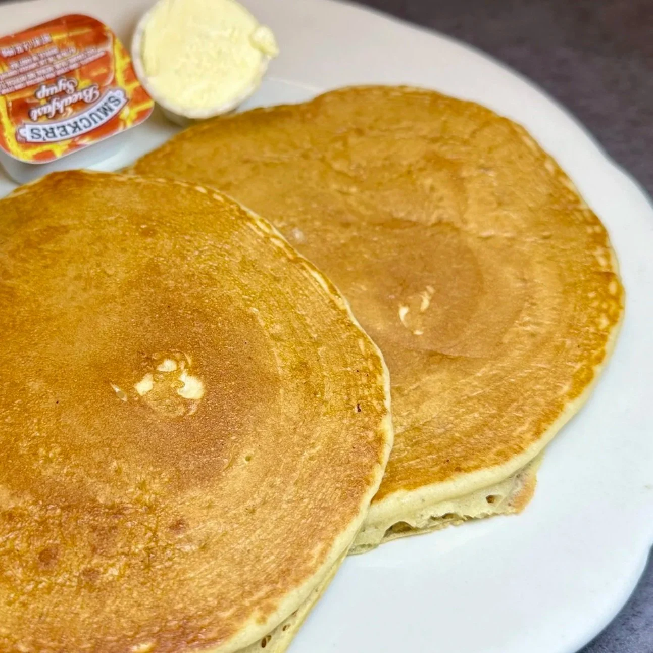 Two golden-brown pancakes on a white plate, with a tub of butter, a container of syrup, and a small dish of butter in the background.