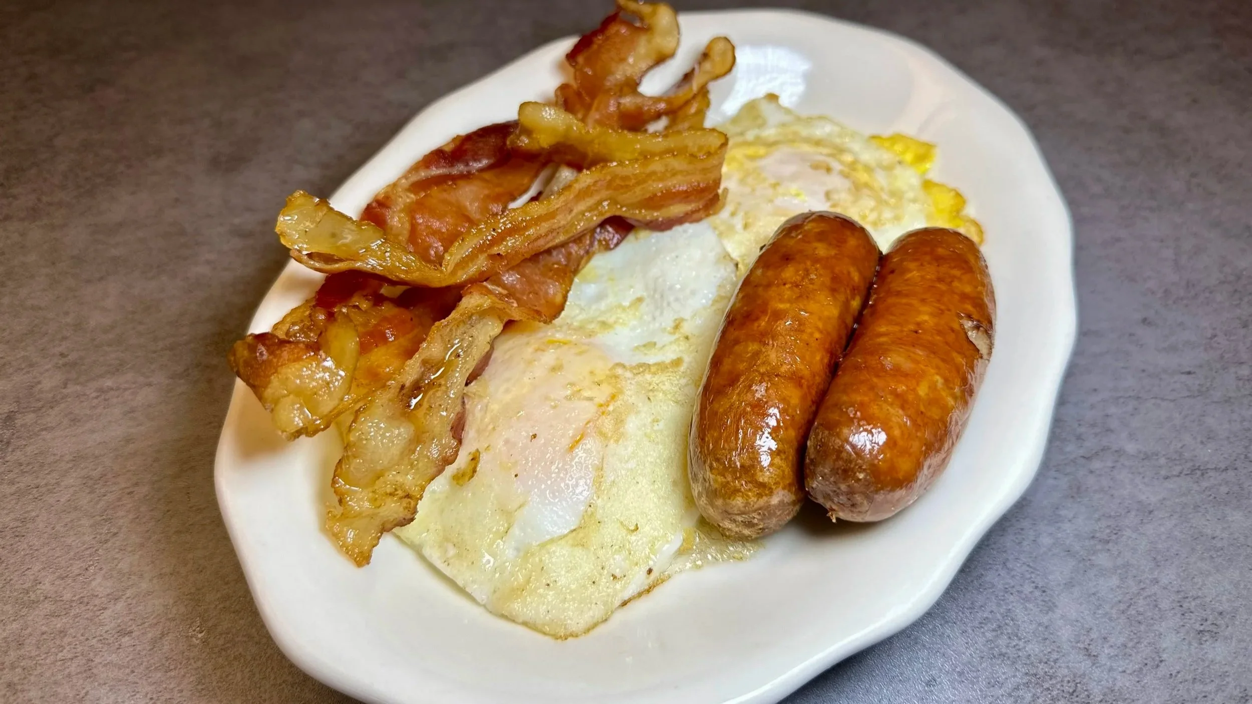 A plate of breakfast food including two cooked eggs, two sausages, crispy bacon, and fried potatoes on a white plate.