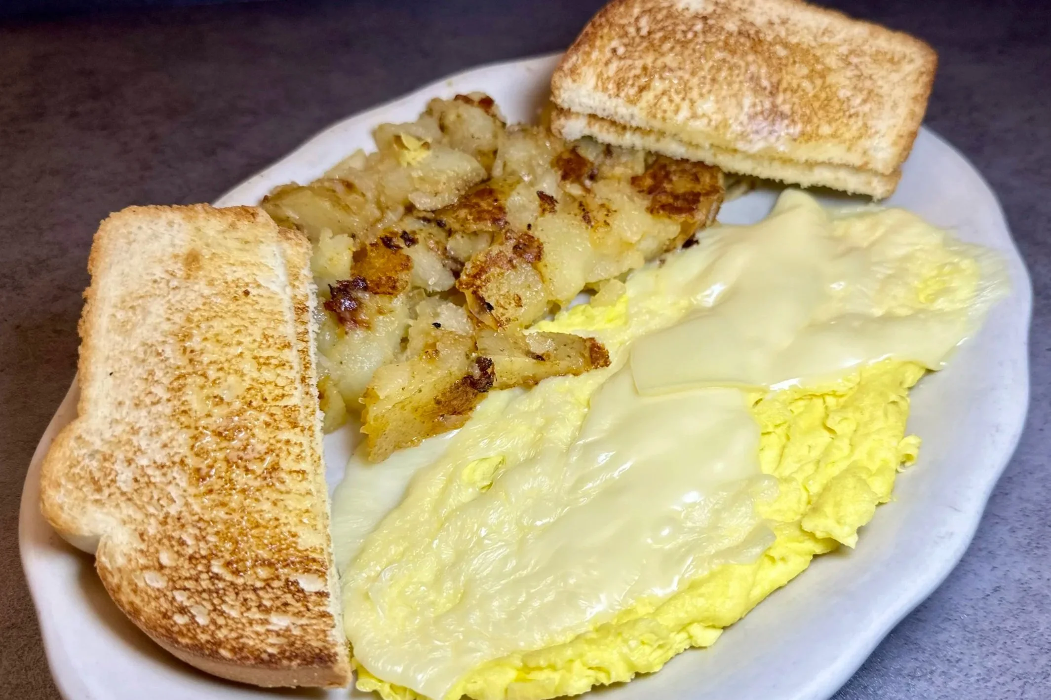 Breakfast plate with scrambled eggs topped with cheese, fried potatoes, and toasted bread slices.