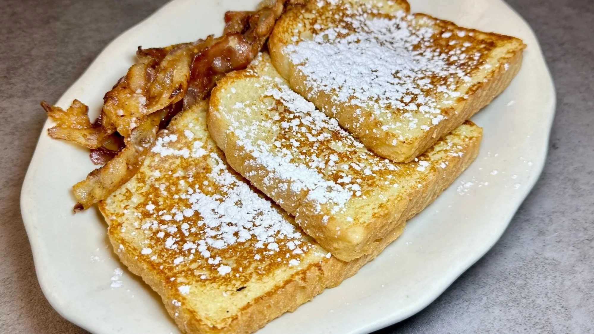 Two slices of French toast topped with powdered sugar, served on a white plate, with bacon on the side.