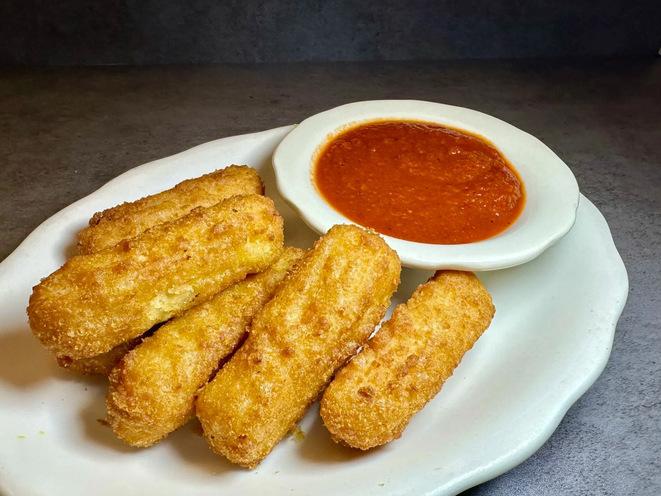 Four breaded fried fish sticks on a white plate with a small bowl of red dipping sauce.