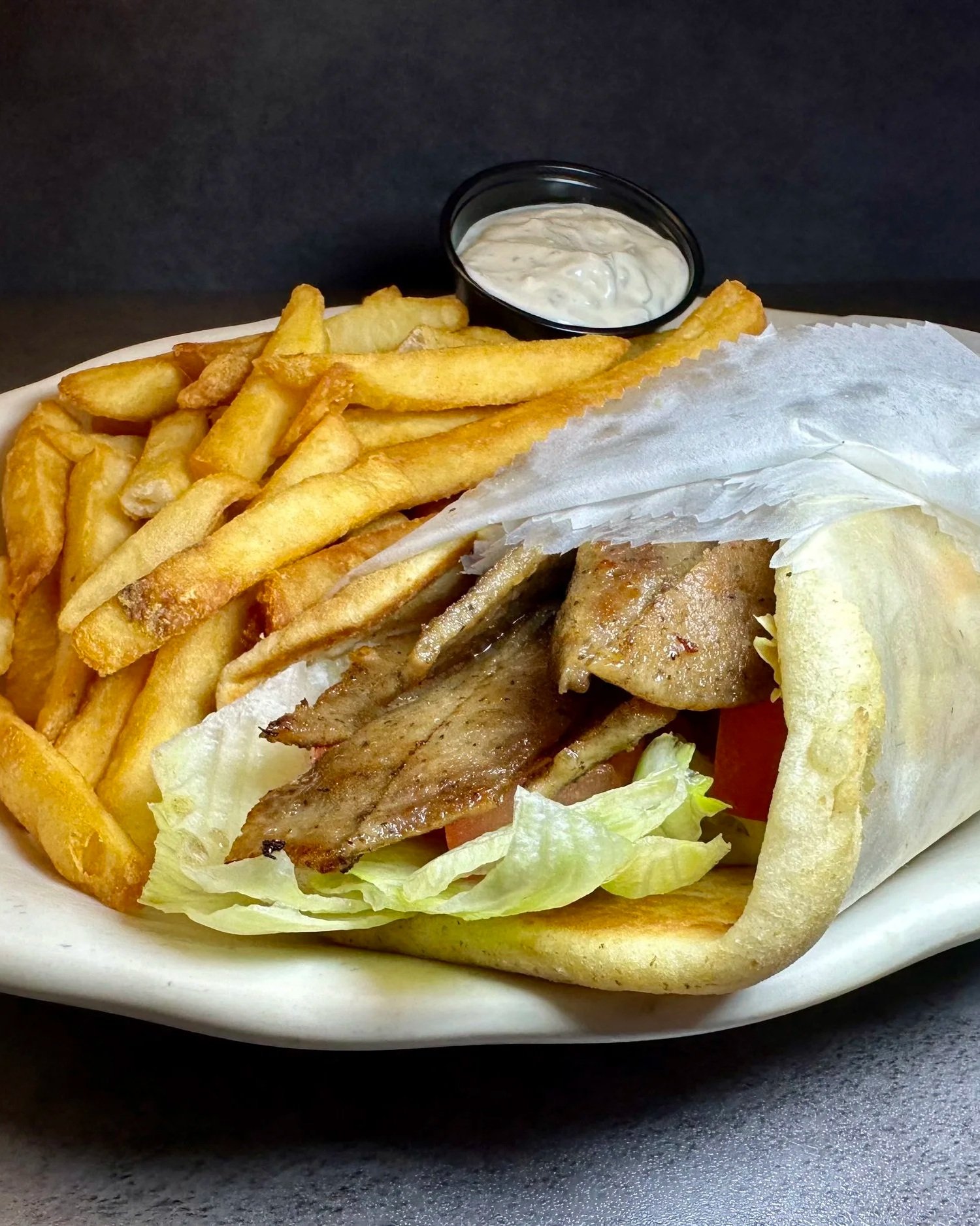 A plate with French fries, a gyro wrapped in pita bread with grilled meat, lettuce, tomato, and a side of creamy sauce, served with a small container of dipping sauce.