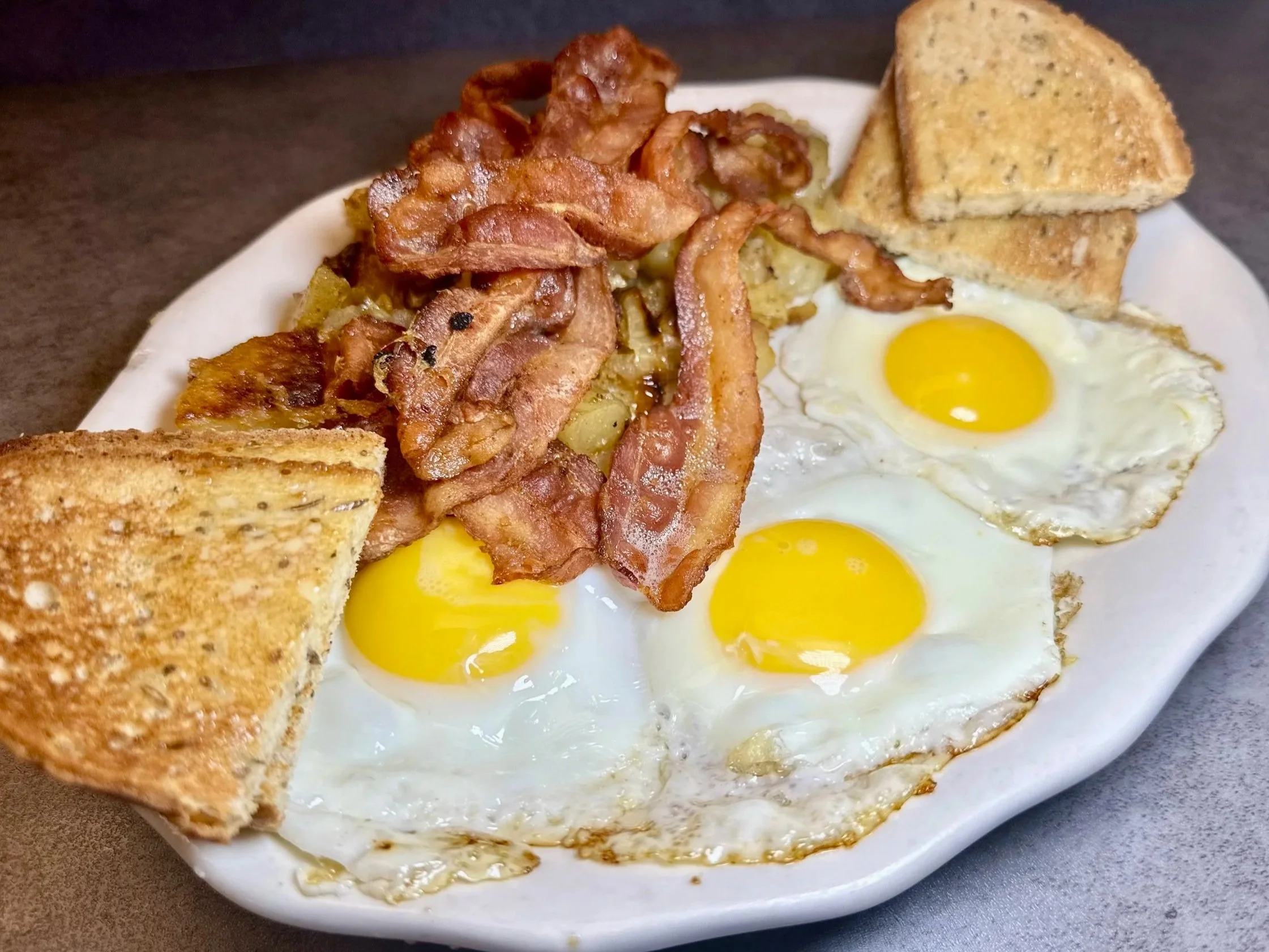 Plate with three fried eggs, crispy bacon, toasted bread triangles, and hash browns.