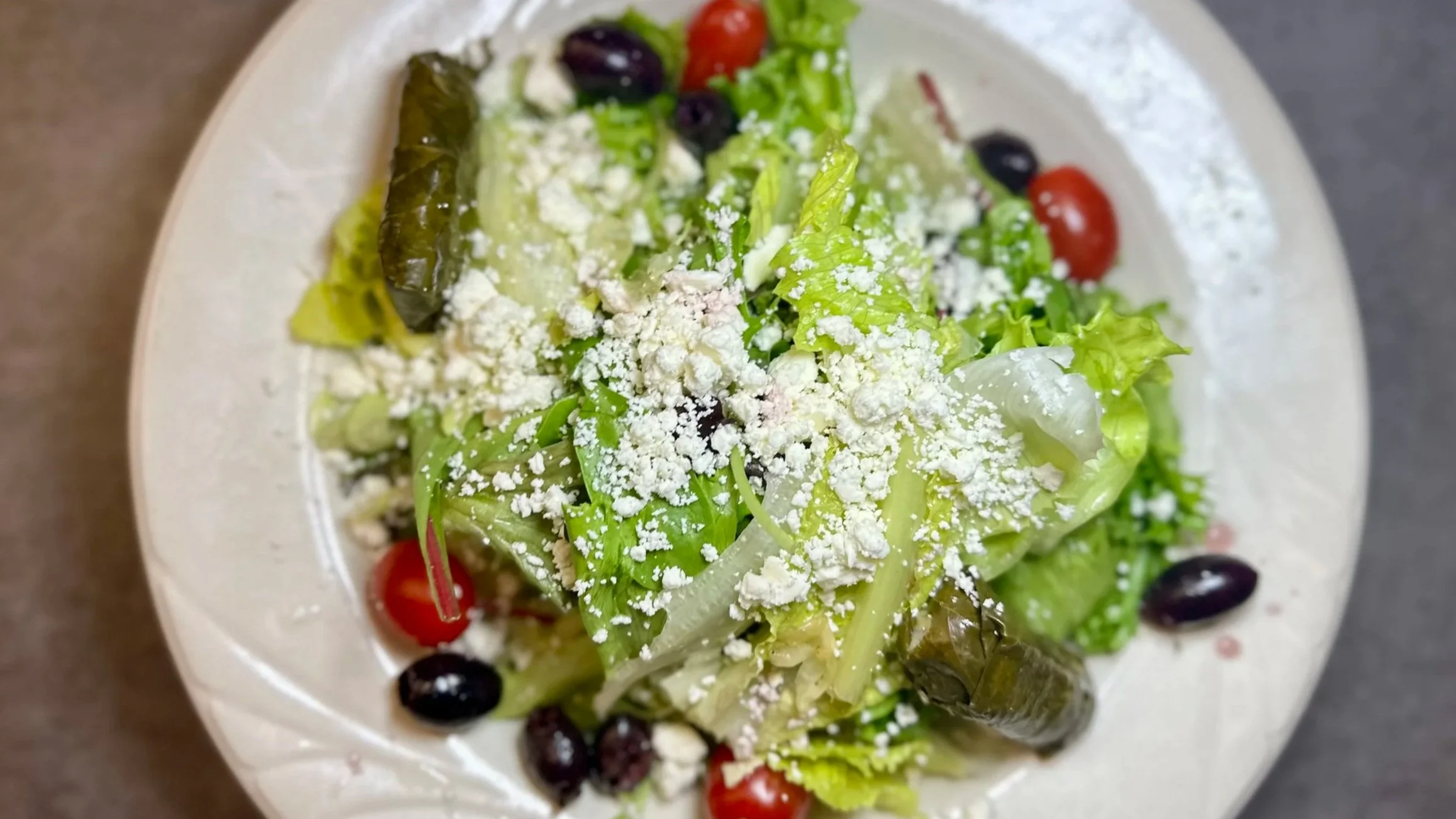 Mixed green salad with cherry tomatoes, black olives, and shredded cheese on a white plate.