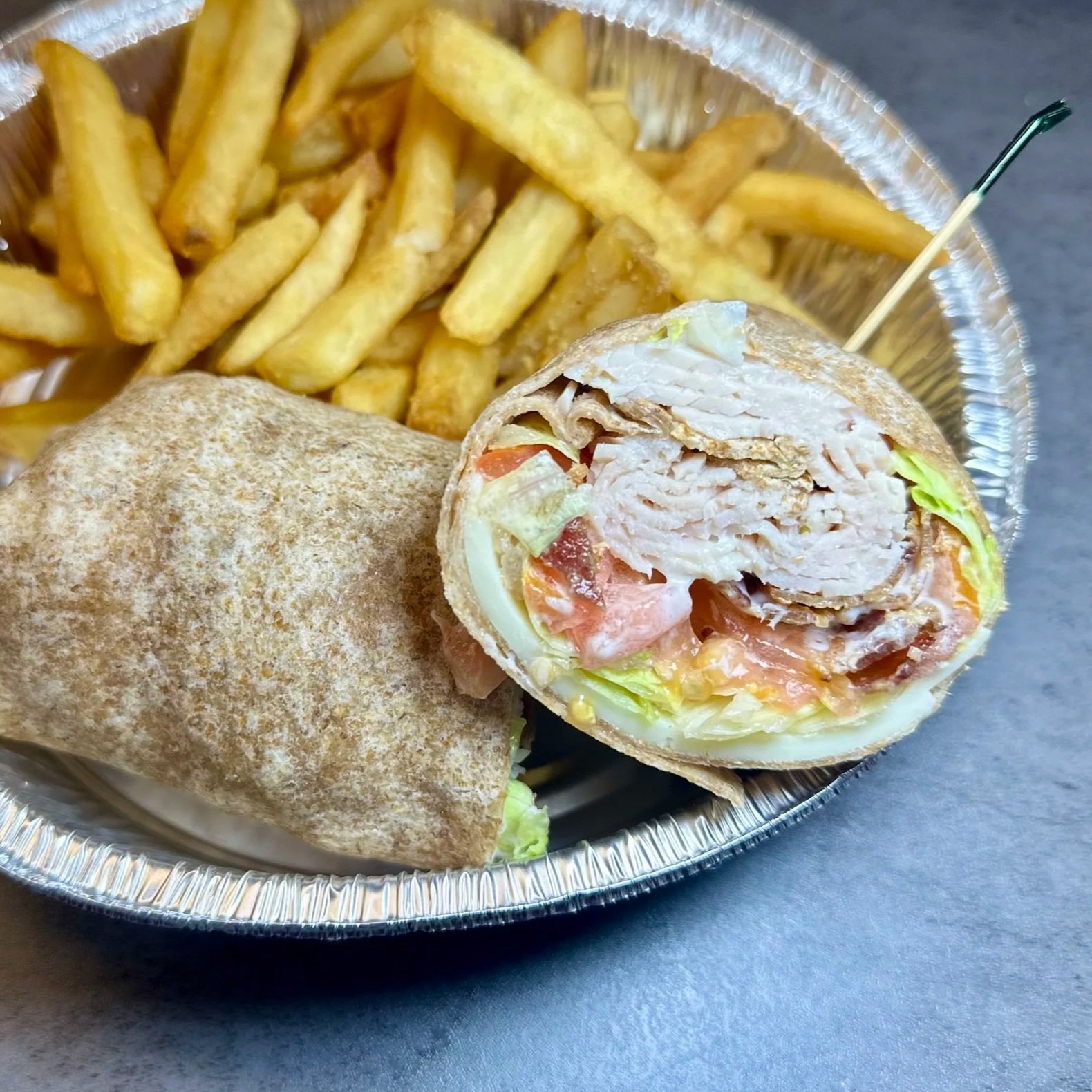 A foil tray containing a chicken wrap with lettuce, tomato, and creamy sauce, alongside a serving of French fries.