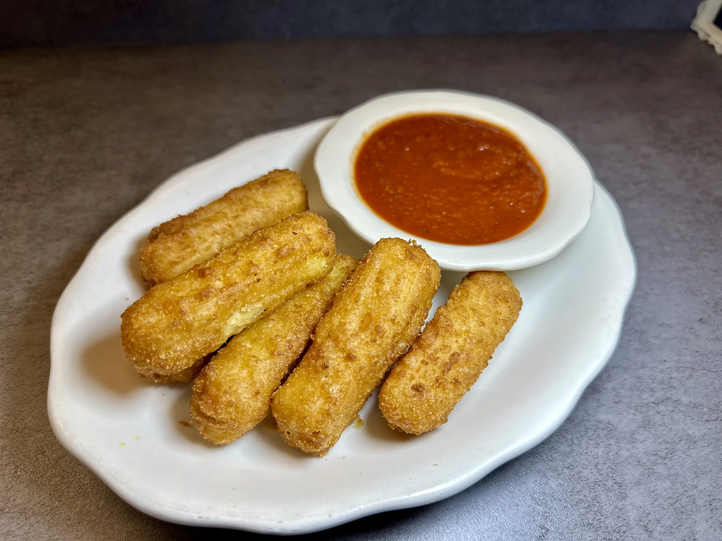 A white plate with five breaded mozzarella sticks and a small bowl of marinara sauce.