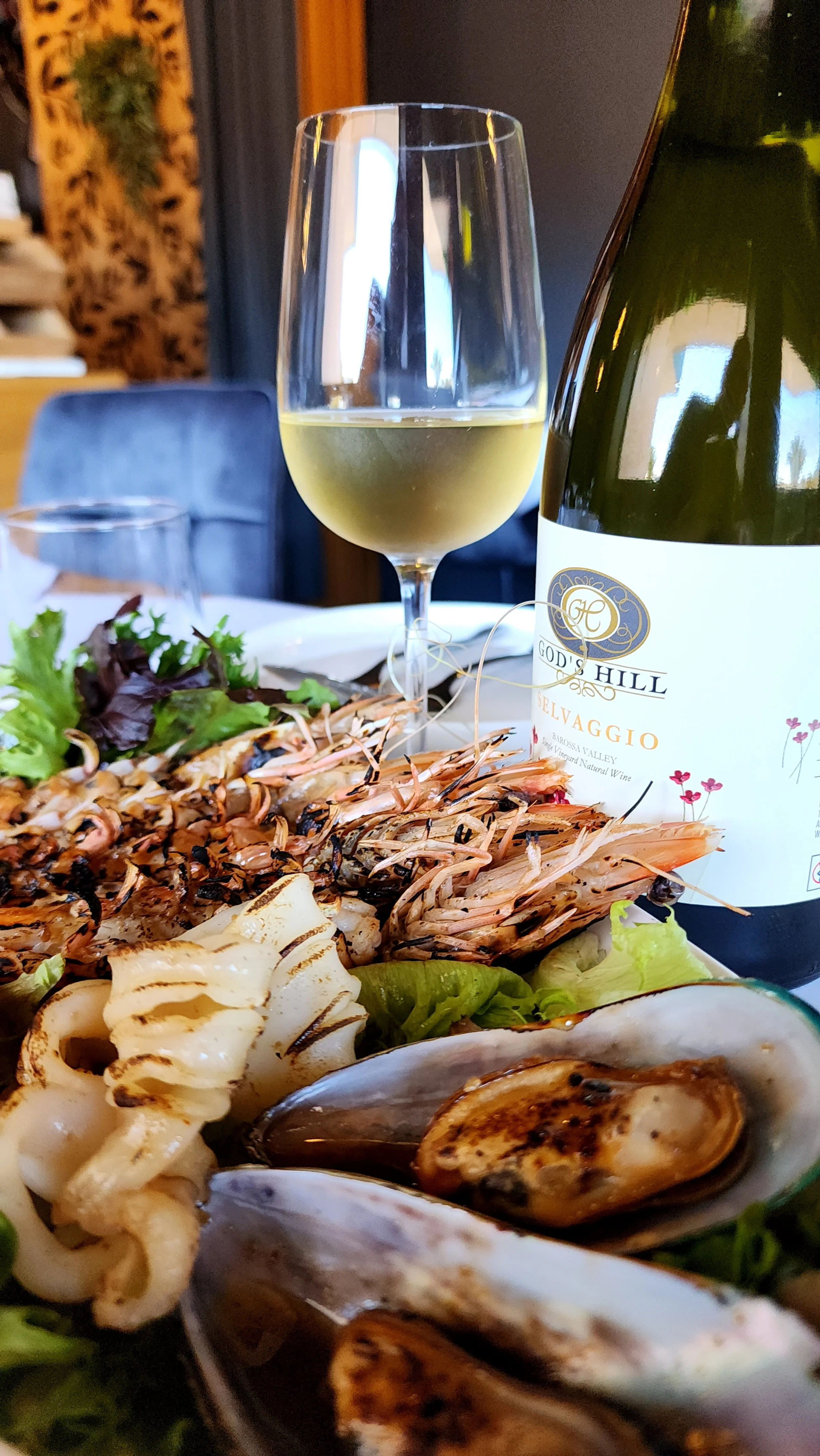 Close-up of a seafood dinner with grilled prawns, mussels, and grilled seafood on a plate, with a glass of white wine and a bottle of wine in the background.