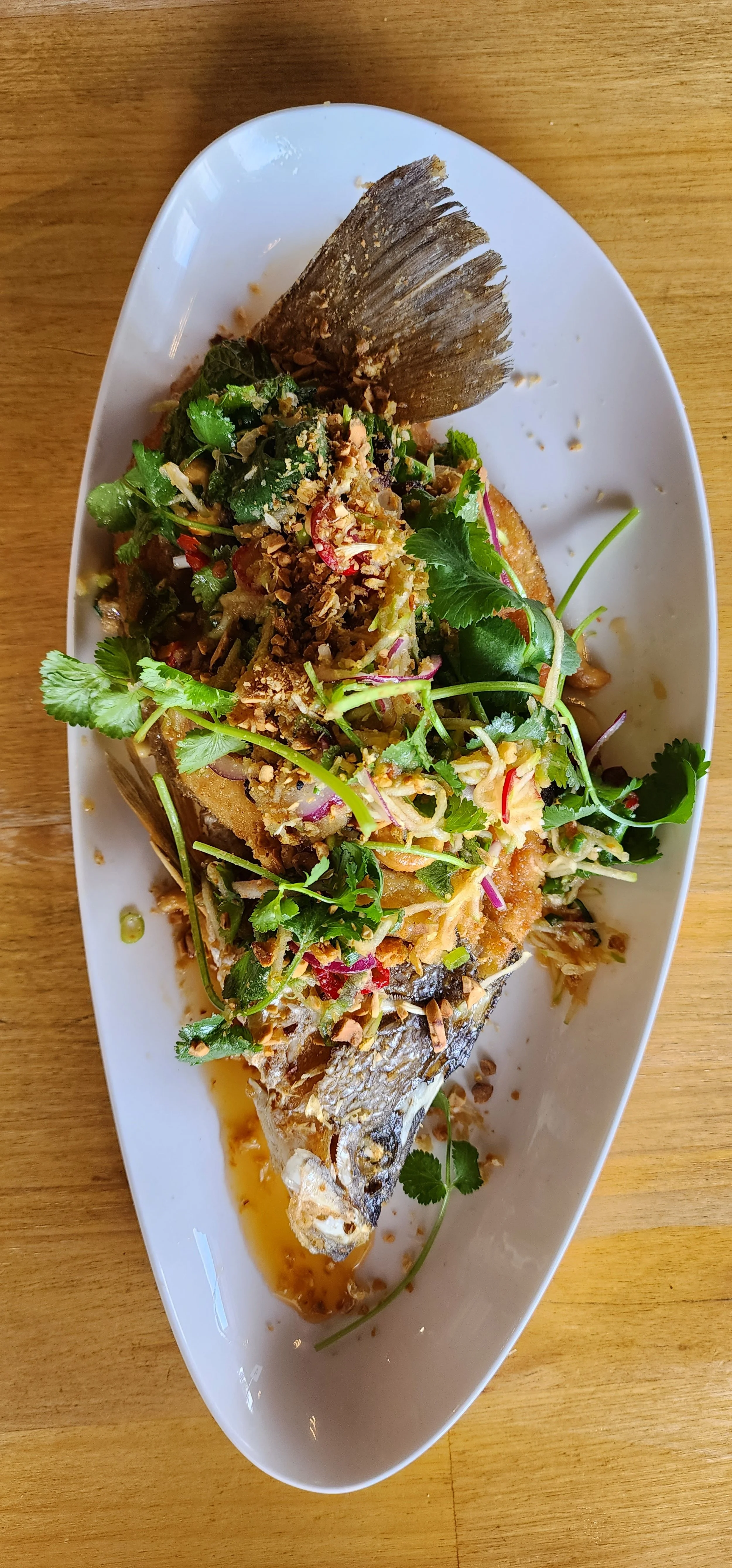 Whole cooked fish topped with herbs, chopped peanuts, and vegetables, served on an oval white plate.