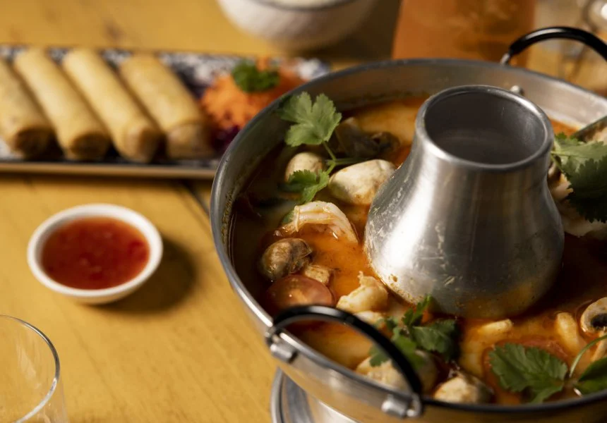 Tom Yum seafood soup with coriander and vegetables, with spring rolls and dipping sauce on a wooden table.